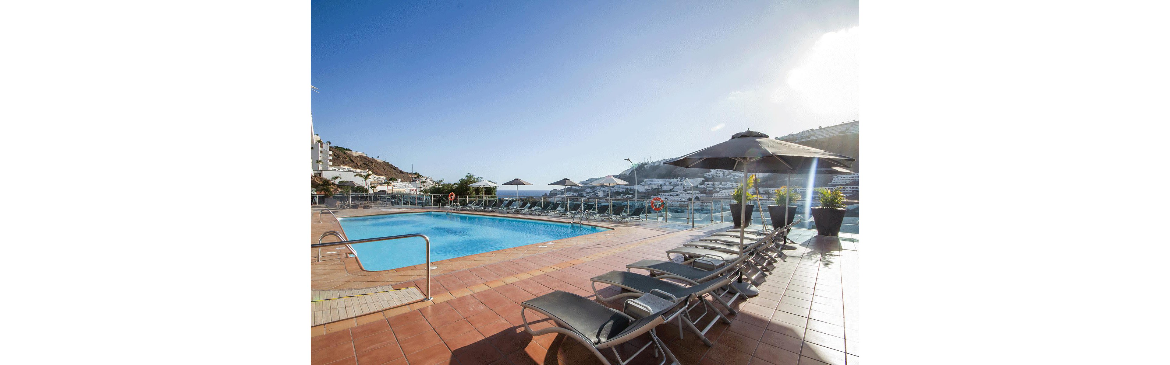 Outdoor swimming pool on a tiled terrace overlooks a hillside resort town and distant sea. Multiple sun loungers with dark umbrellas line the poolside, suggesting a relaxing vacation setting. Clear blue sky and bright sunlight create a warm, inviting atmosphere. Glass safety railings, potted plants, and a lifebuoy add detail to the modern hotel environment.