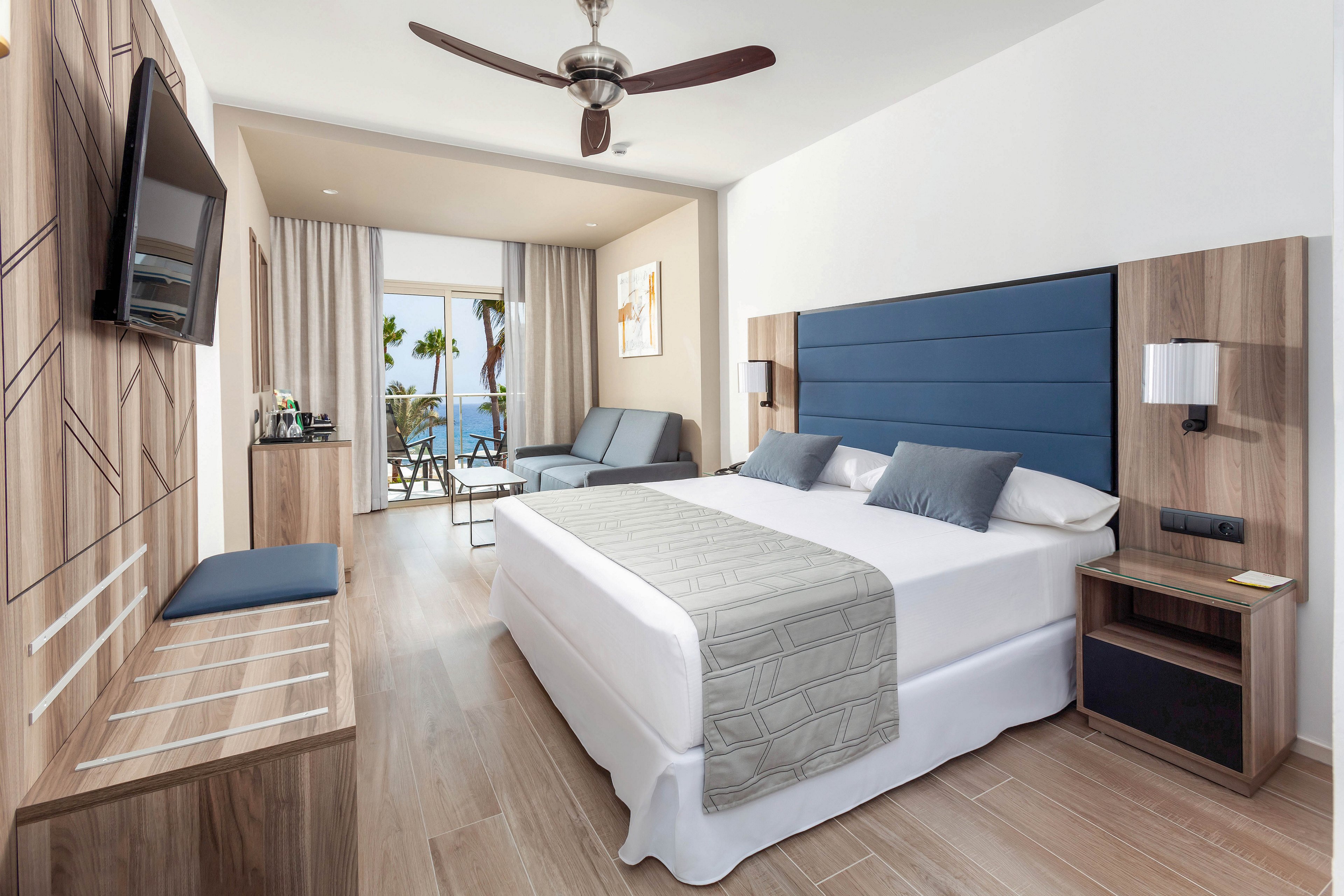 A contemporary hotel room features a large bed with blue and gray accents, a wall-mounted TV, and a seating area with a sofa. The room opens to a balcony with a table and chairs, offering a view of palm trees and the ocean. The decor is minimalist with neutral tones and natural light fills the space.