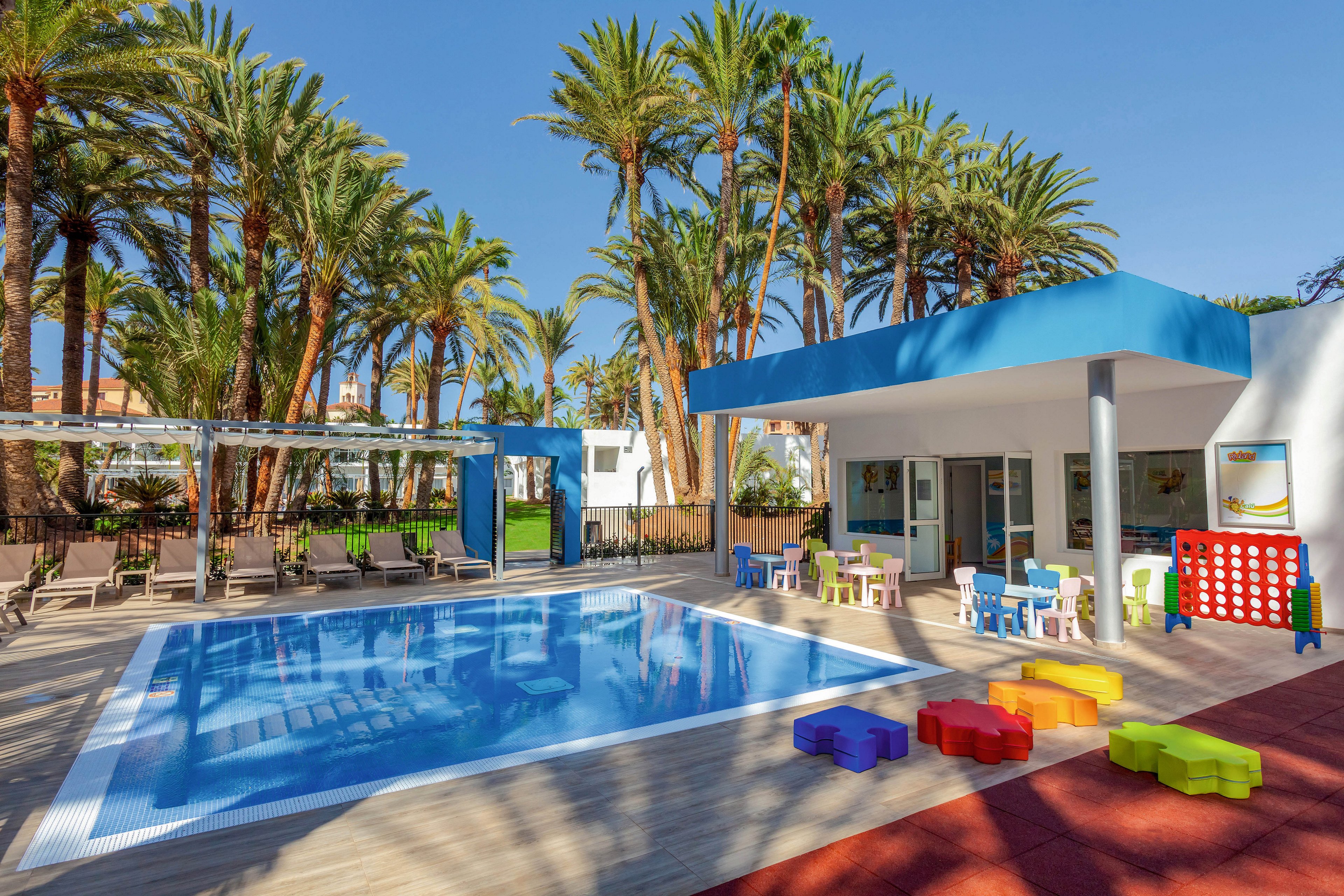 A modern outdoor swimming pool is surrounded by palm trees and sun loungers, creating a relaxing resort atmosphere. A shaded area features colorful children's furniture and large outdoor games, indicating a family-friendly environment. The scene is bright and sunny, with a playful mood and vibrant color palette.