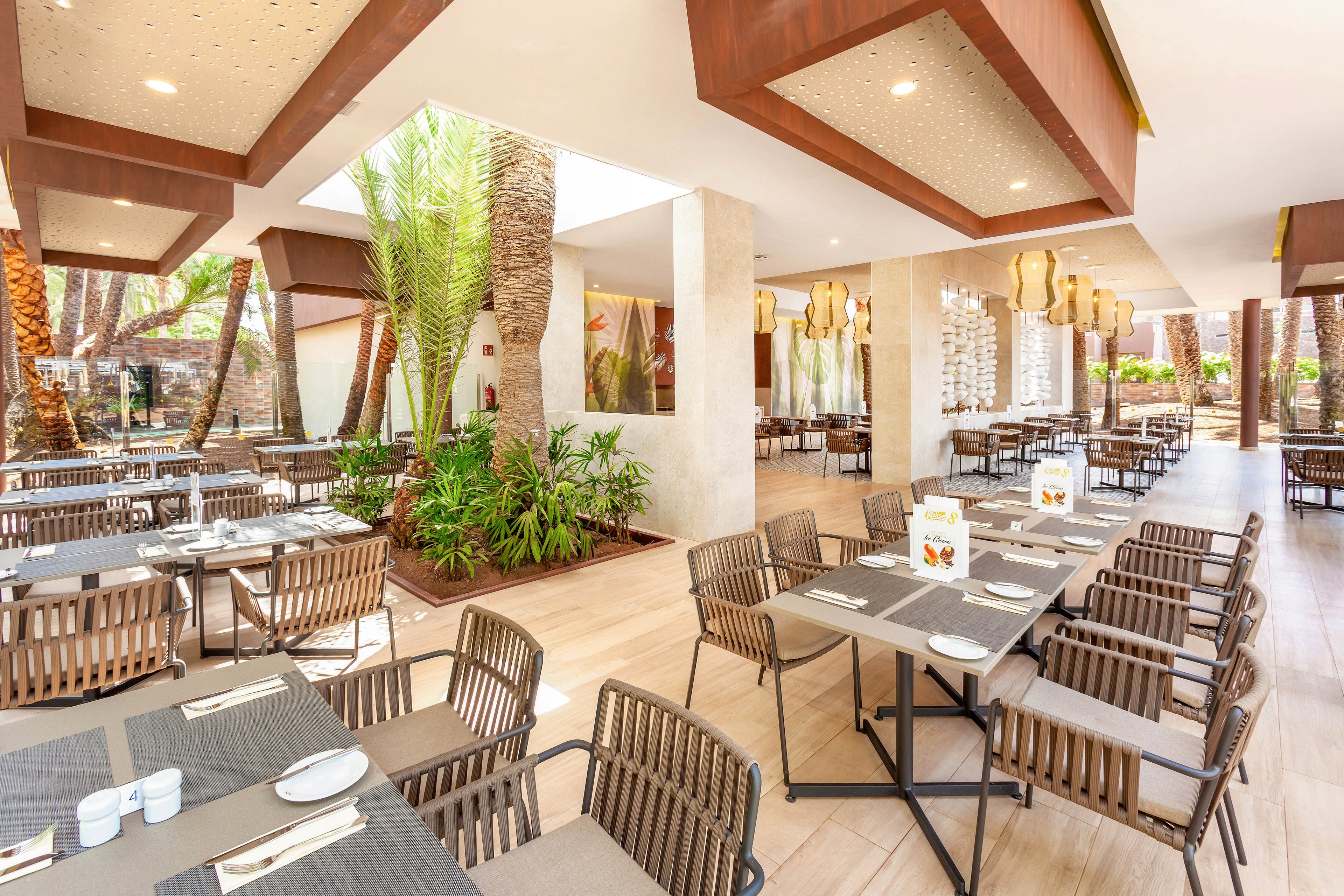 A spacious, contemporary restaurant features neatly arranged tables and chairs in an open-air setting. The dining area is accented by lush indoor plants, wooden decor, and ample natural light streaming through large windows. Table settings include plates, cups, and utensils, with some tables displaying menus or table cards. The overall atmosphere is bright, inviting, and relaxed, with a neutral color palette and modern design elements.