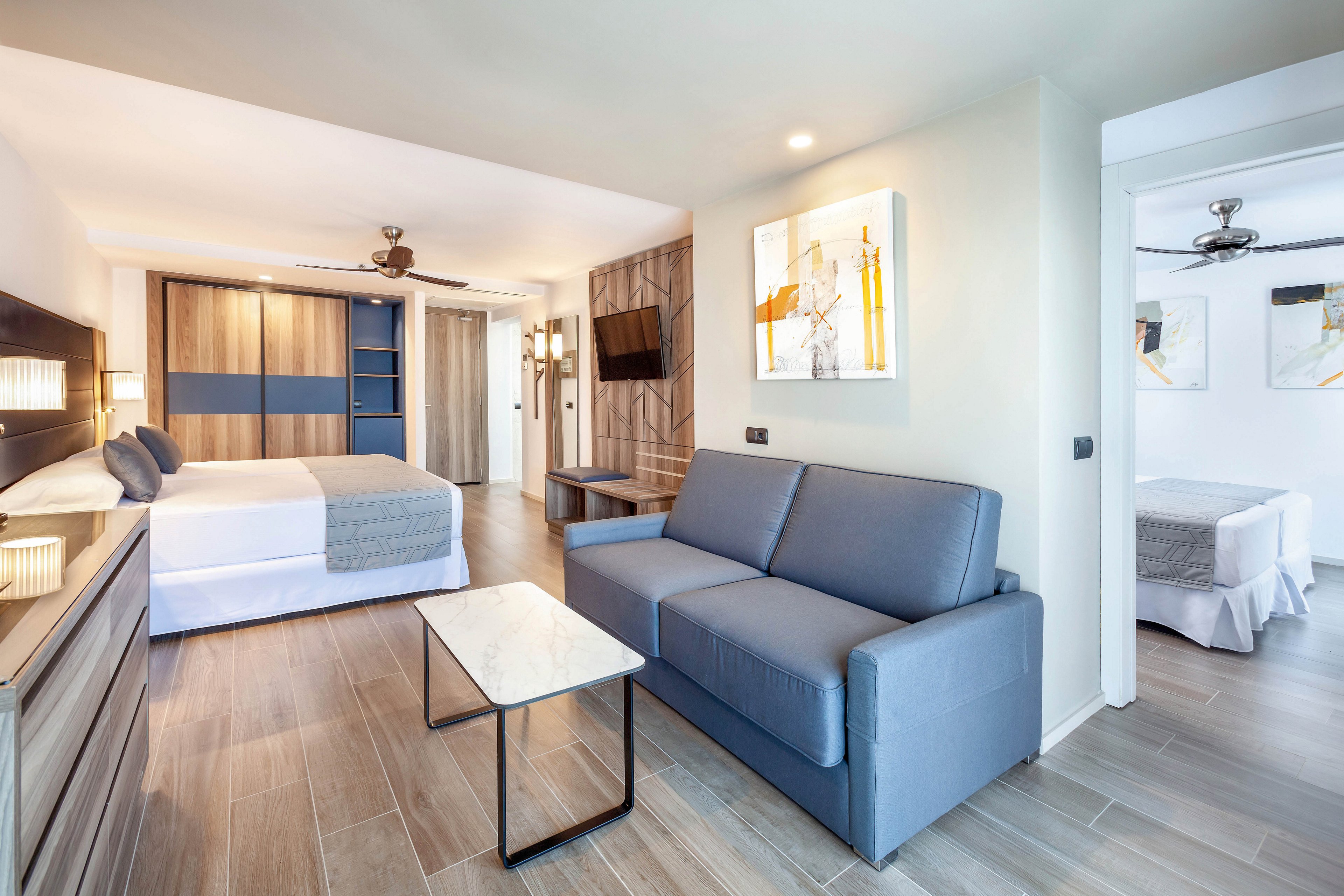 A spacious hotel suite features a contemporary bedroom with a large bed, a stylish lounge area with a blue sofa, and a marble-topped coffee table. The room is decorated in neutral tones with wood accents and abstract artwork on the walls. Natural light fills the space, creating a bright and inviting atmosphere, while ceiling fans add comfort.
