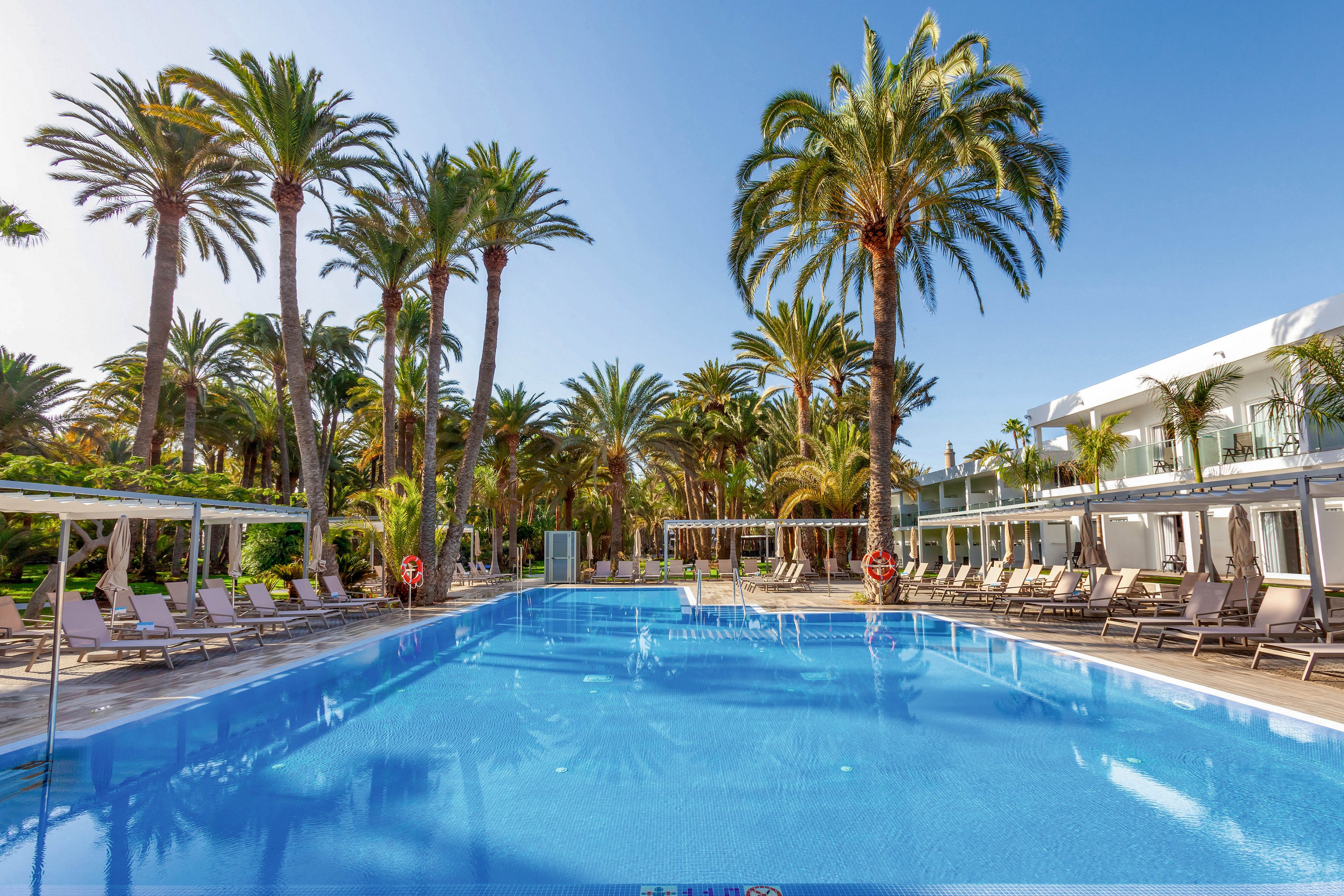 The main subject is a large, inviting swimming pool set in an outdoor resort environment. Lounge chairs line both sides of the pool, shaded by tall palm trees and modern white cabanas. The scene is bright and tranquil, with clear blue skies and reflections on the water creating a relaxing mood. No visible text or numeric values are present in the image.