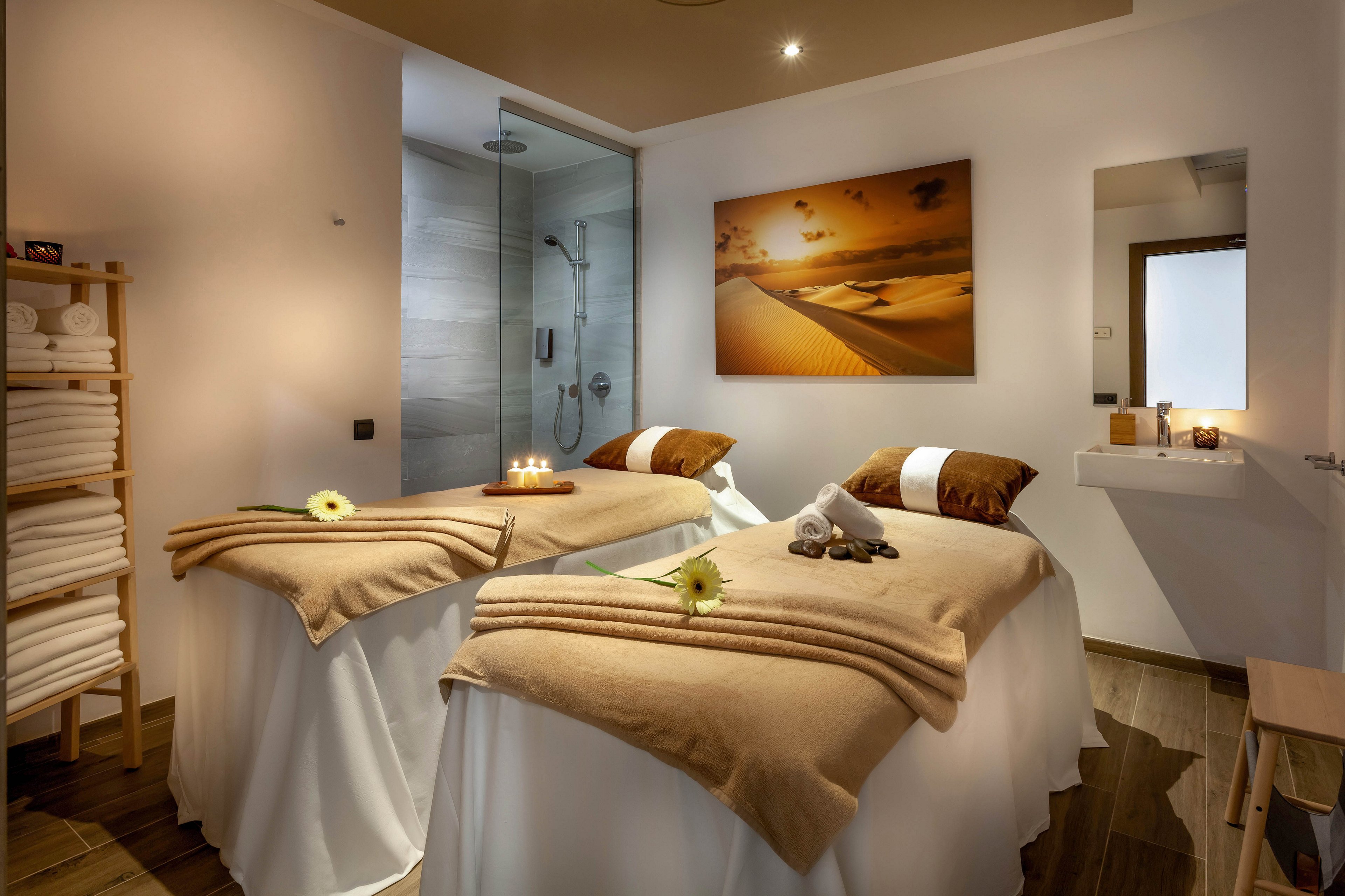A contemporary spa room features two massage tables adorned with beige blankets and brown pillows, each topped with a single flower. The setting includes a shower area, neatly stacked towels, and soft candlelight, creating a tranquil and inviting atmosphere. Wall art depicting a serene desert scene enhances the relaxing mood, while the neutral color palette adds warmth and calm.
