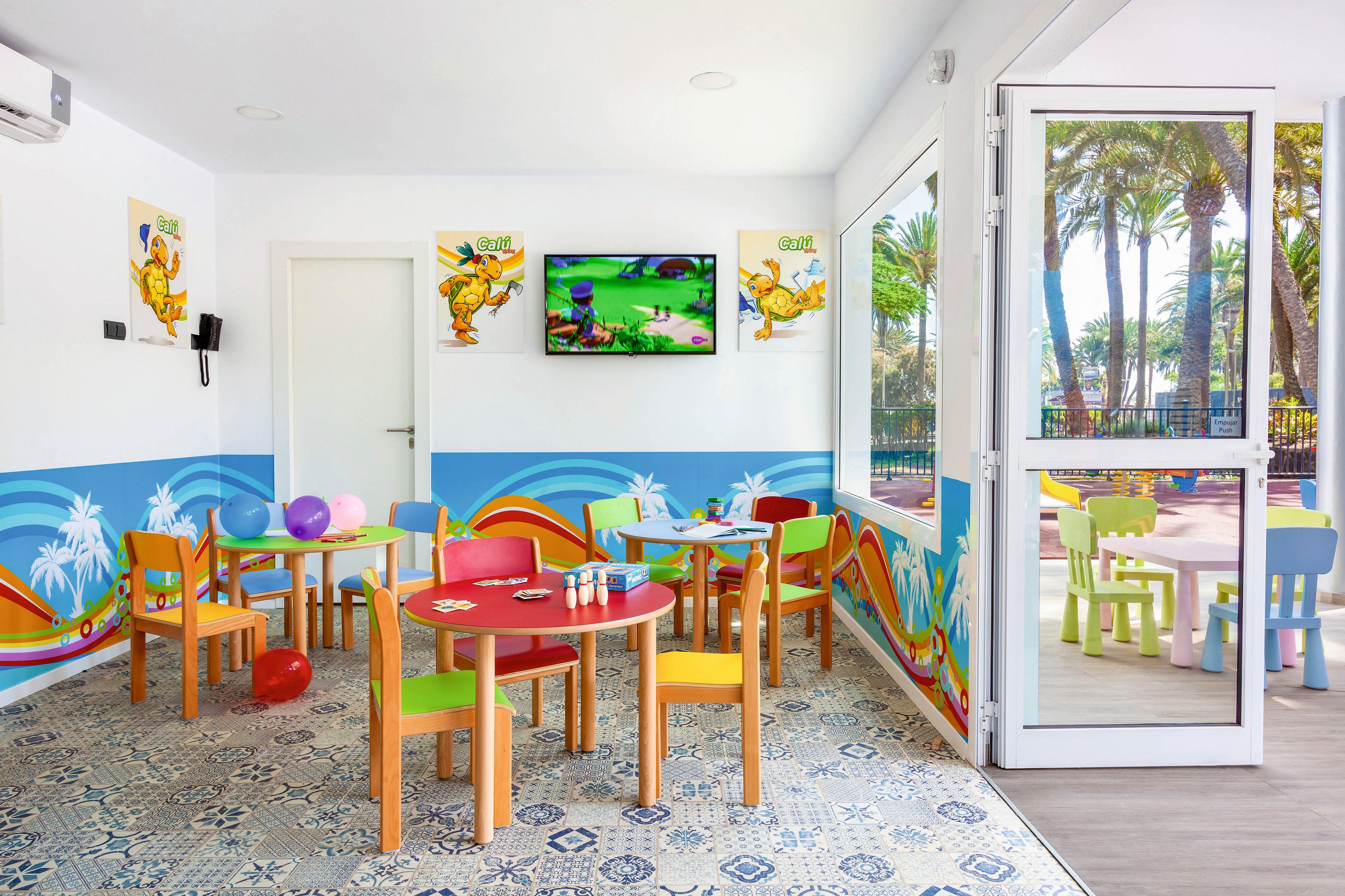 A vibrant children’s playroom features small wooden tables and chairs in various colors. The room is decorated with playful wall art, balloons, and a television displaying a cartoon scene. Large windows and an open door reveal an outdoor area with more child-sized furniture and palm trees, creating a cheerful and inviting atmosphere.