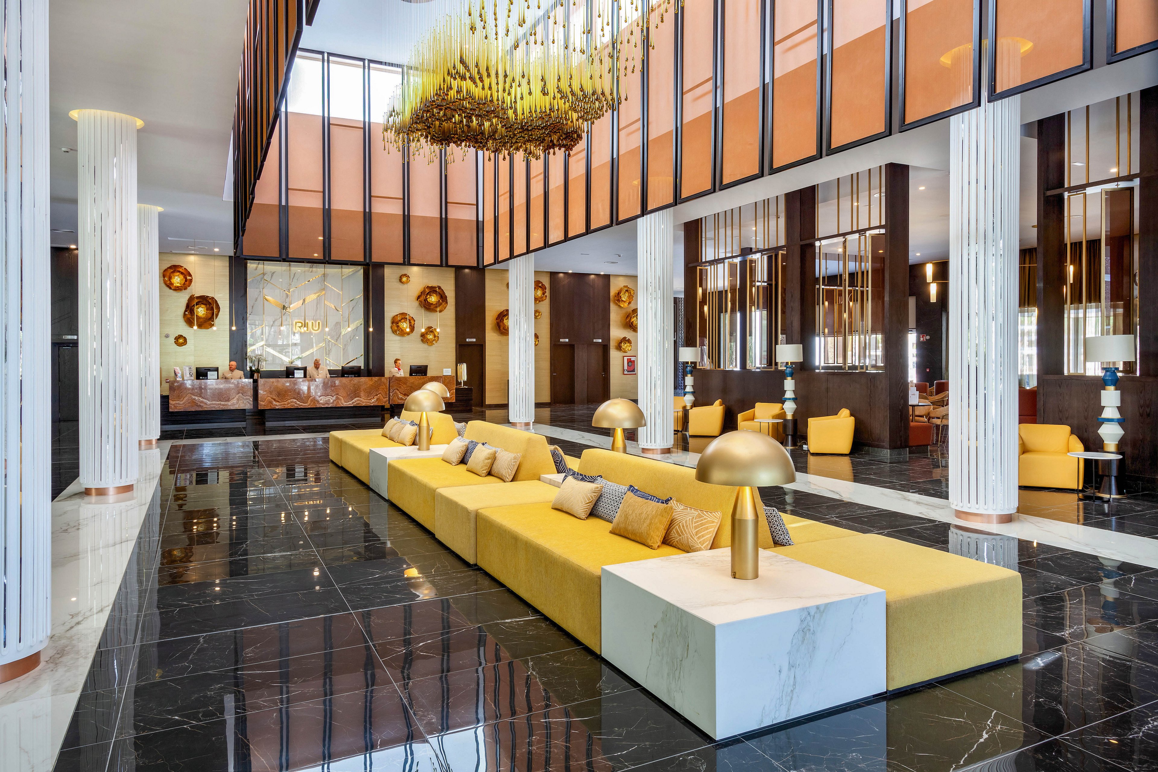 A spacious, contemporary hotel lobby features long yellow sofas with marble side tables and gold lamps. The setting is bright and elegant, with high ceilings, large windows, and a dramatic gold chandelier as a focal point. Decorative wall art and reflective surfaces enhance the luxurious atmosphere. The color palette includes black, white, gold, and yellow tones. No visible text or numbers are present in the image.