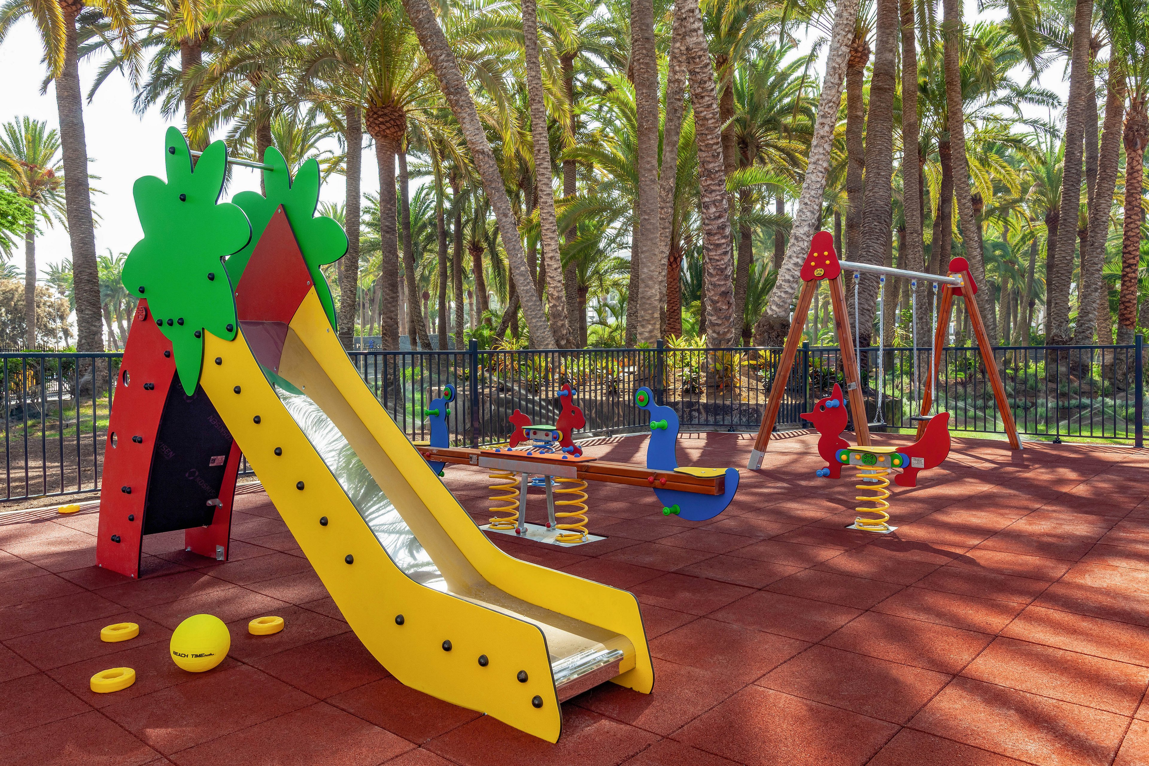 A vibrant children's playground features a yellow and red slide shaped like a tree, spring riders, and a swing set. The play area is set on a rubberized surface surrounded by tall palm trees and a black fence. Bright sunlight and lush greenery create a cheerful, inviting atmosphere. A yellow ball and several play pieces are scattered on the ground. No visible text or numbers are present in the image.