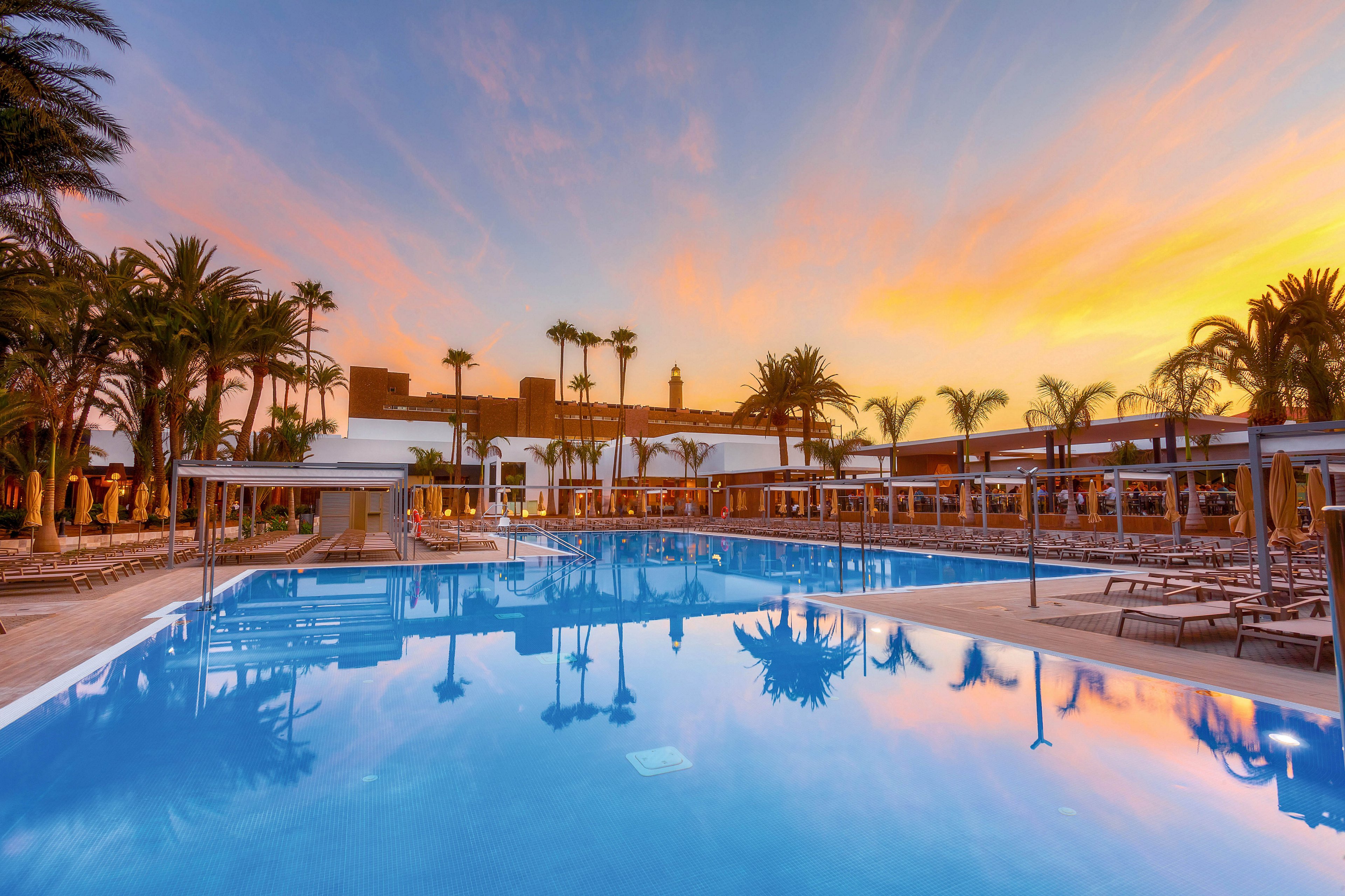 A large outdoor swimming pool is the main focus, surrounded by lounge chairs and palm trees. The setting is a resort, captured during a vibrant sunset with warm orange and pink hues reflecting on the water. The tranquil atmosphere and clear sky enhance the relaxing mood, with no visible people or text in the image.