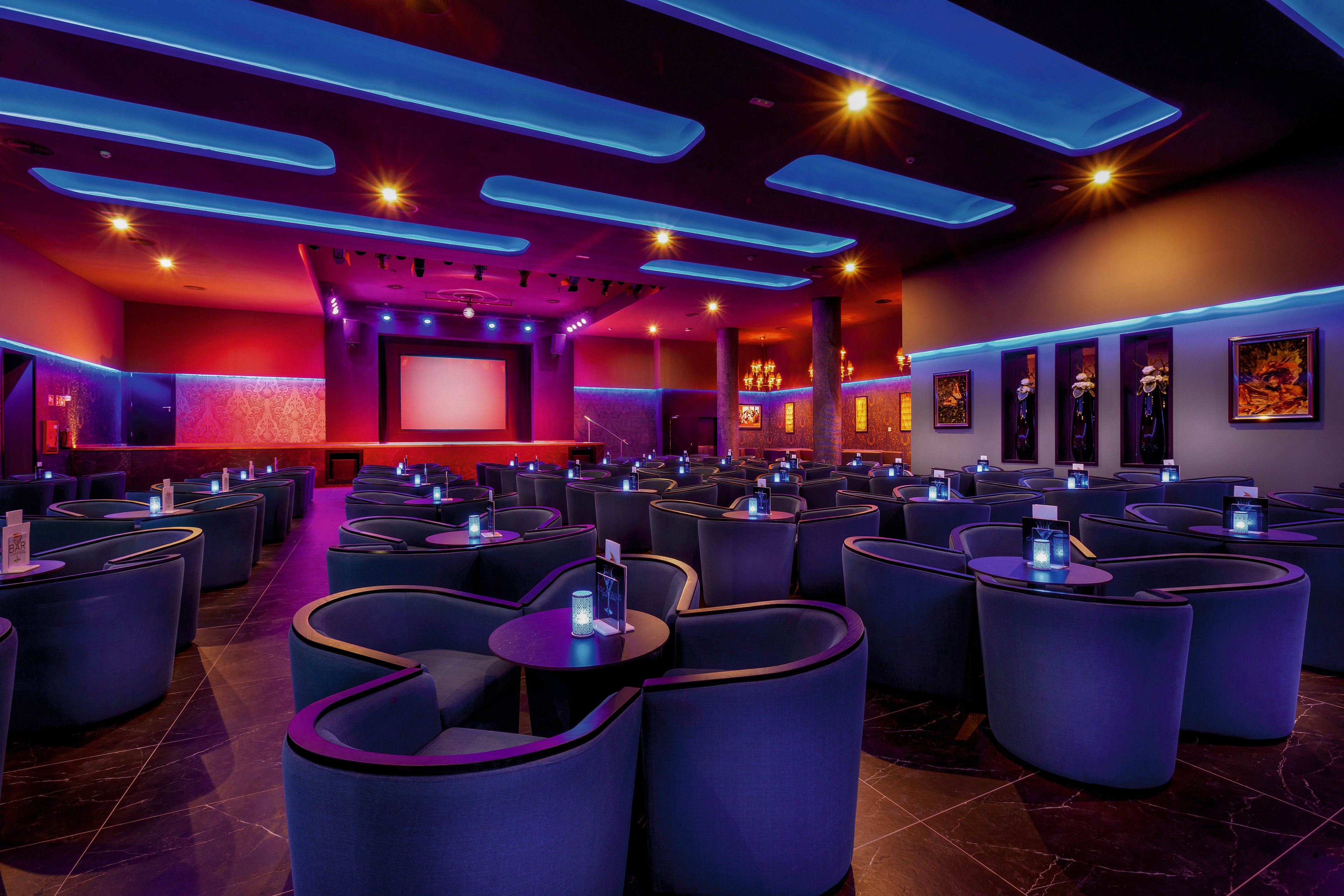 The image shows a stylish cinema lounge featuring curved armchairs and round tables arranged in rows. The setting is illuminated with vibrant blue and purple LED lights, creating a contemporary and inviting atmosphere. A large projection screen is visible at the front, with decorative wall art and glowing fixtures enhancing the mood. No visible text or numeric values are present in the image.