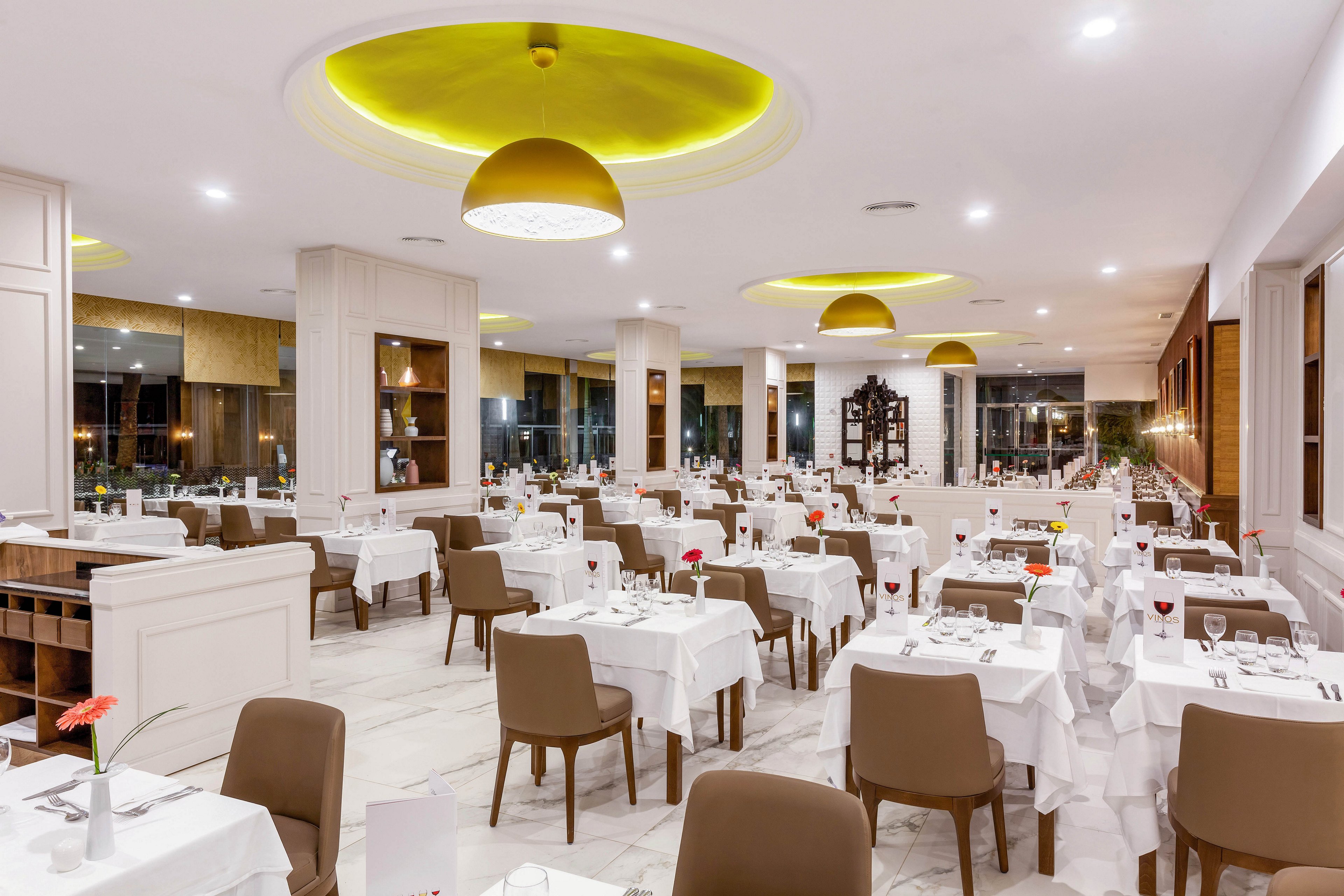 The image shows a spacious, contemporary restaurant dining area with neatly arranged tables covered in white linens. Brown chairs complement the clean, minimalist decor, and yellow ceiling lights add warmth to the atmosphere. The setting is empty, emphasizing the elegant and inviting ambiance. No visible text or numeric values are present in the scene.