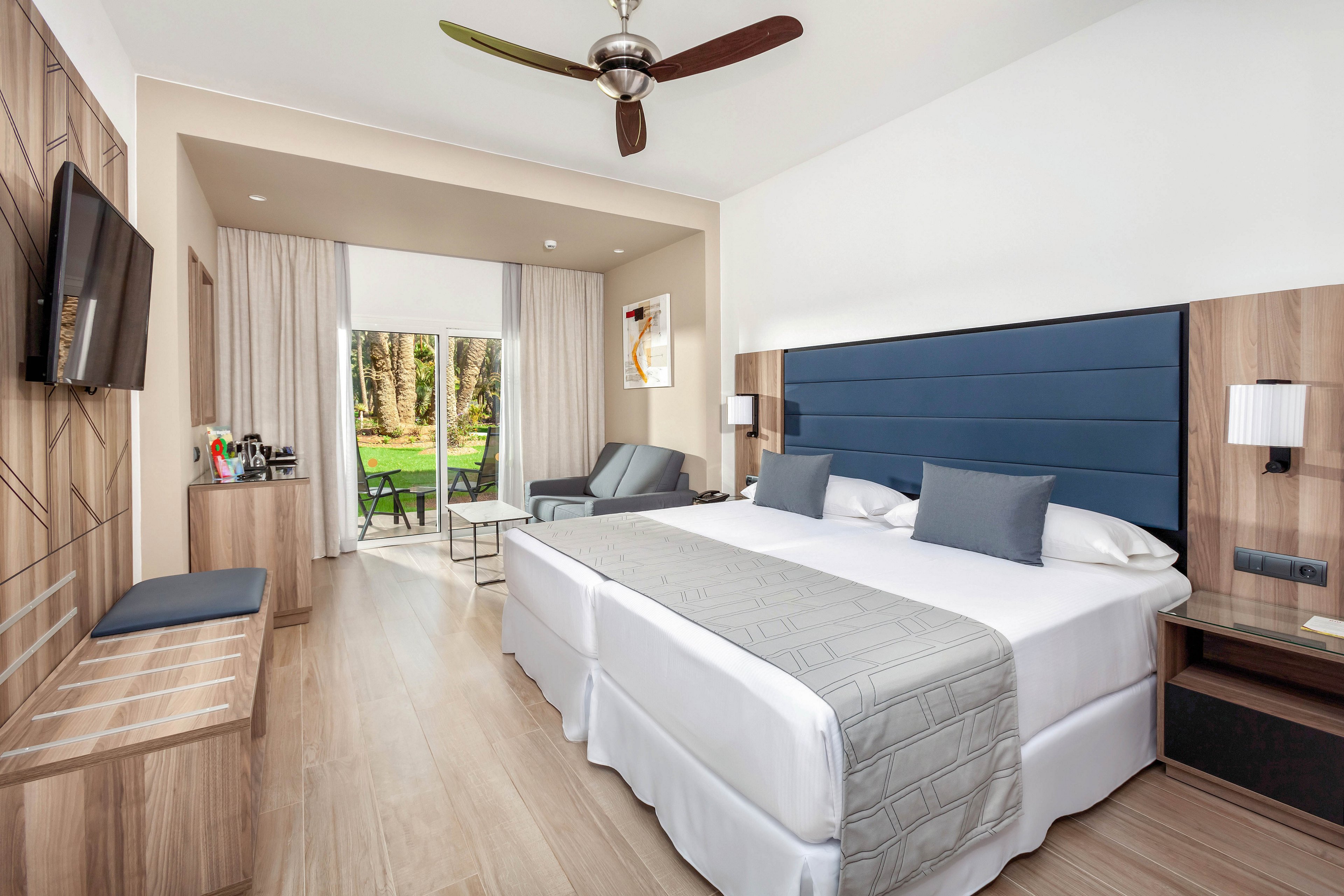 A spacious, contemporary hotel room features a large king-size bed with gray and white bedding. The room includes a flat-screen TV, a sitting area with a sofa, a desk, and a coffee station. Large windows open to a lush garden view, and the decor is minimalist with neutral tones and wood accents. No visible text or numbers are present in the image.