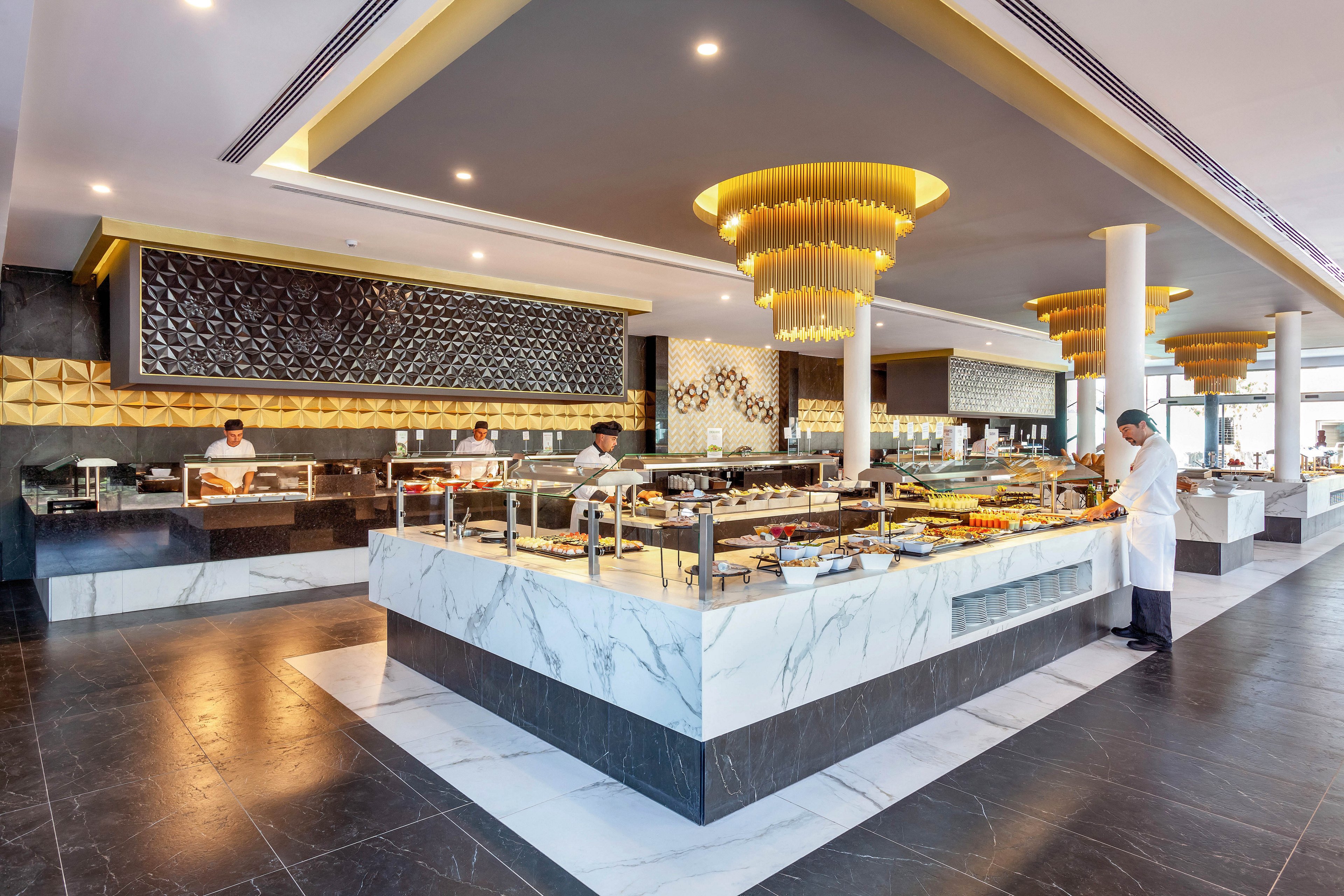 A spacious hotel buffet features marble serving counters and elegant gold lighting fixtures. Several chefs in white uniforms are preparing and presenting food behind the counters. The setting is contemporary with geometric wall accents and a clean, open layout. The mood is professional and inviting, with a neutral and gold color palette.