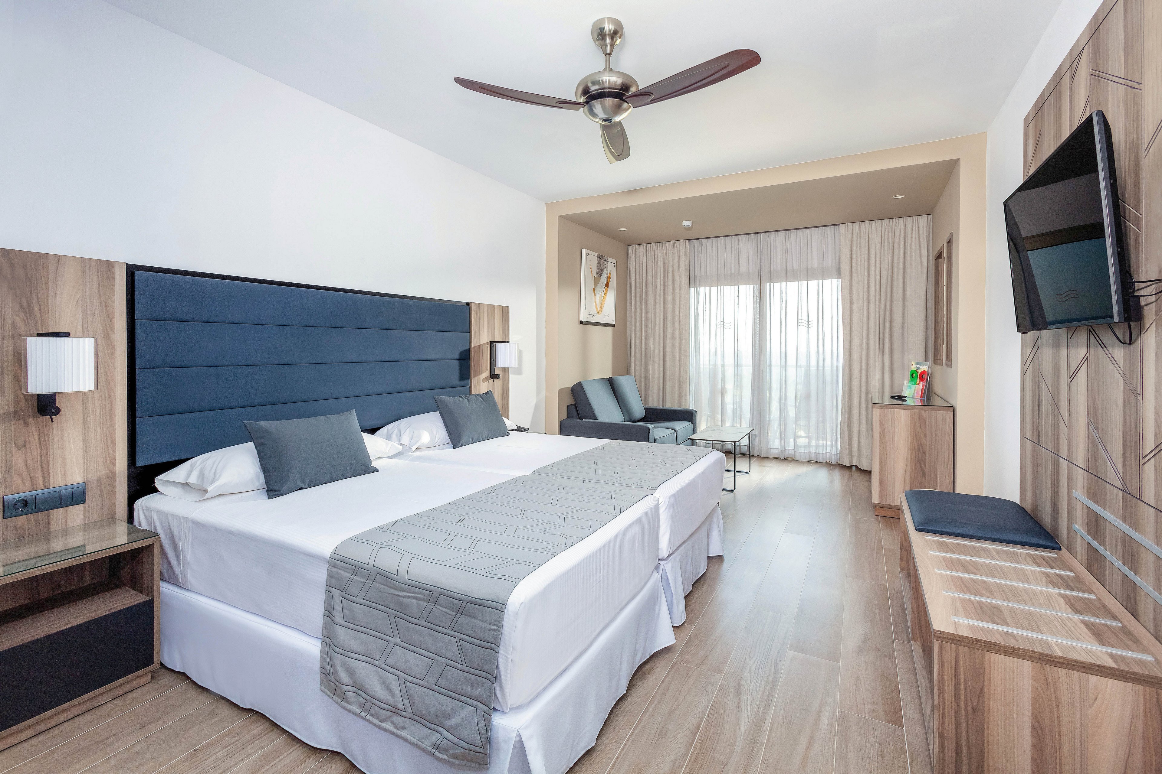 A contemporary hotel room features two twin beds with gray headboards and matching bedding. The space includes a wall-mounted television, a ceiling fan, and a sitting area with a sofa near large windows leading to a balcony. The room is decorated in neutral tones with wood accents, creating a calm and inviting atmosphere.