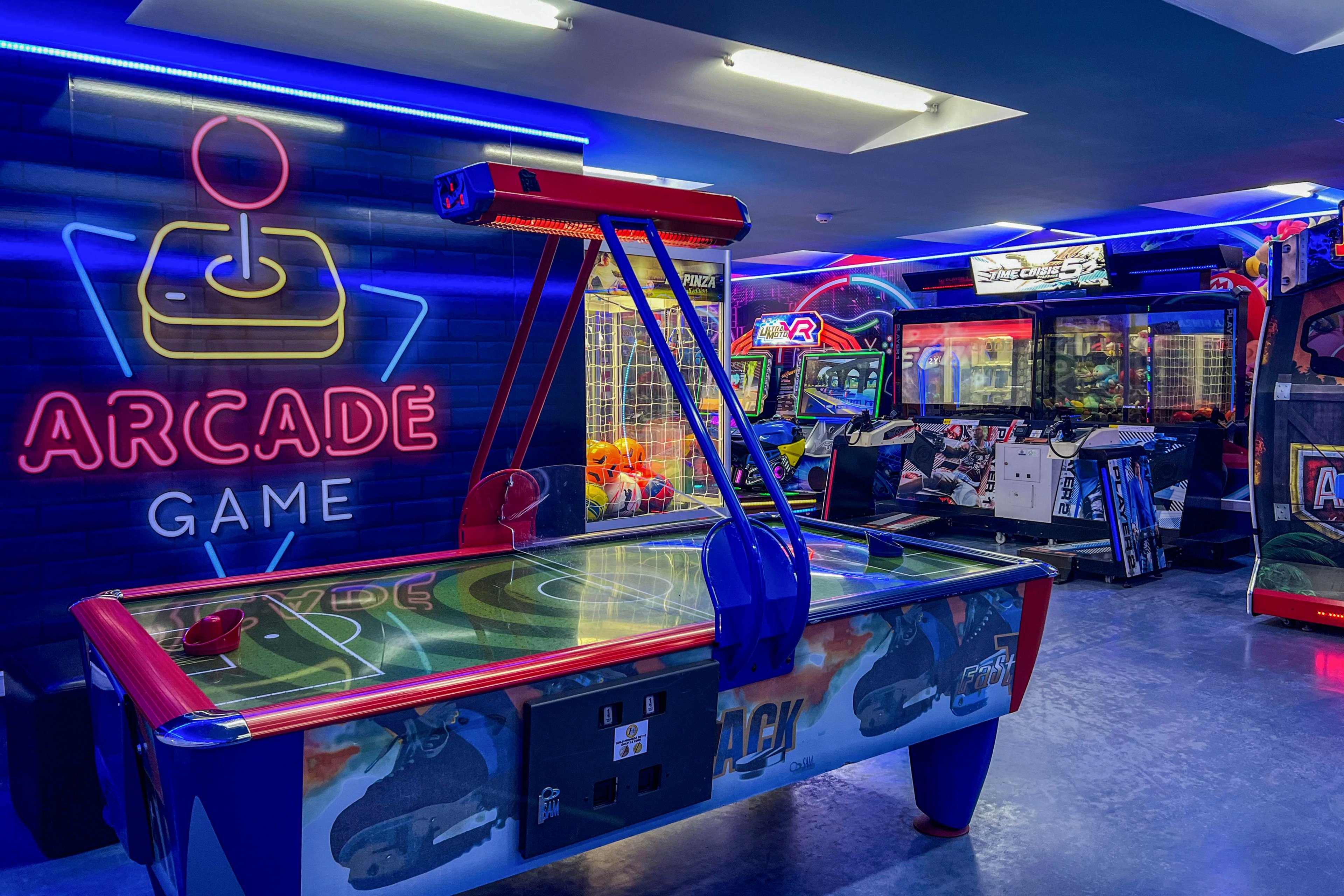 A brightly lit arcade room features a prominent air hockey table in the foreground. Behind it, various arcade machines and claw games line the walls, creating a lively entertainment setting. Blue and pink neon lights illuminate the space, including a large wall graphic with a joystick icon and the words ARCADE GAME. The overall atmosphere is playful and futuristic, emphasizing fun and leisure.
