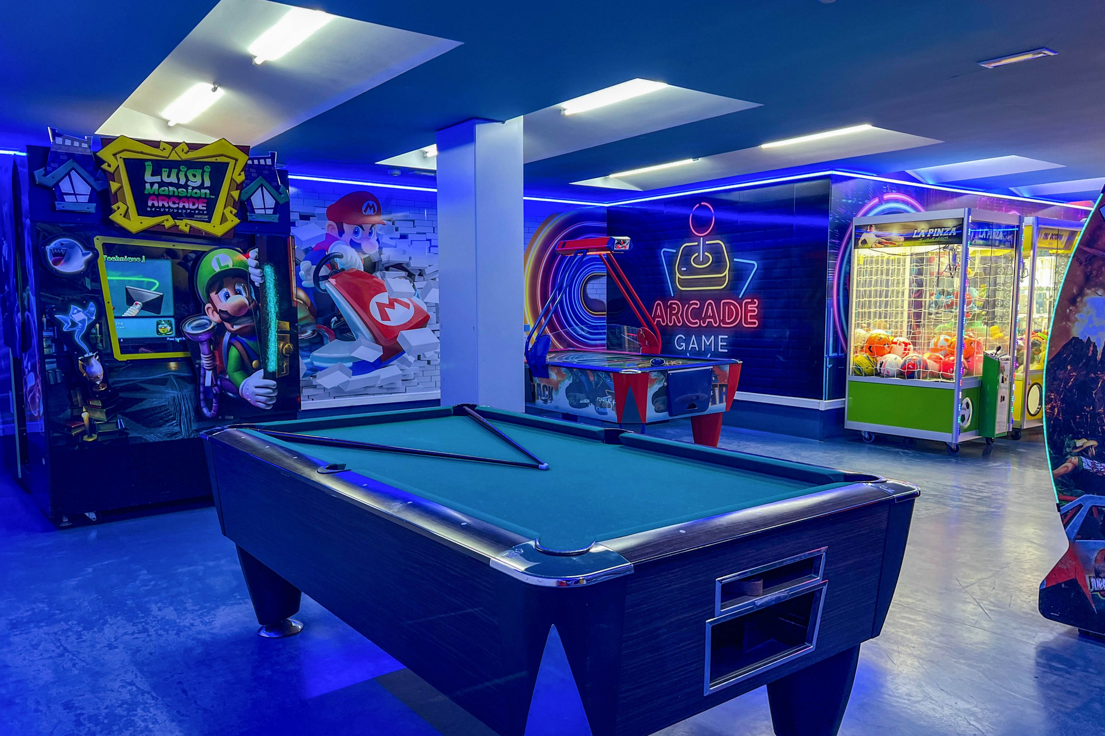 A modern arcade room features a central pool table under bright ceiling lights. Along the walls are colorful arcade machines, including a racing game and claw machines, all glowing with blue neon accents. Large wall graphics and the words "ARCADE GAME" add to the retro gaming atmosphere. The space appears empty, emphasizing the vibrant machines and illuminated environment.