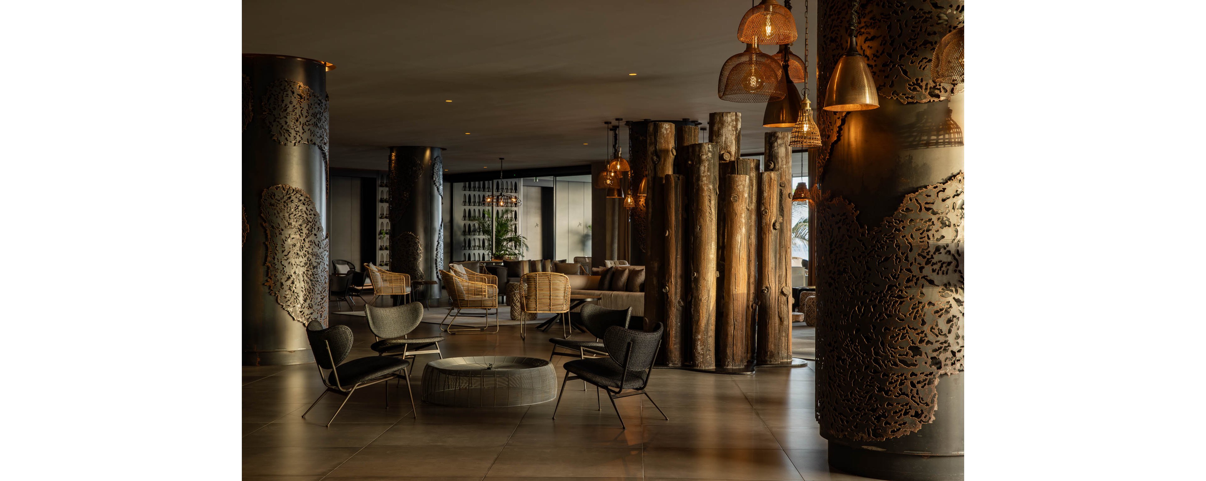A spacious hotel lobby features modern lounge chairs and woven armchairs arranged around low tables. Large sculptural wooden columns and carved metal pillars create a striking focal point in the center of the room. Pendant lights with warm amber tones hang from the ceiling, casting a soft, inviting glow. Floor-to-ceiling windows in the background reveal greenery and add natural light to the sophisticated interior.