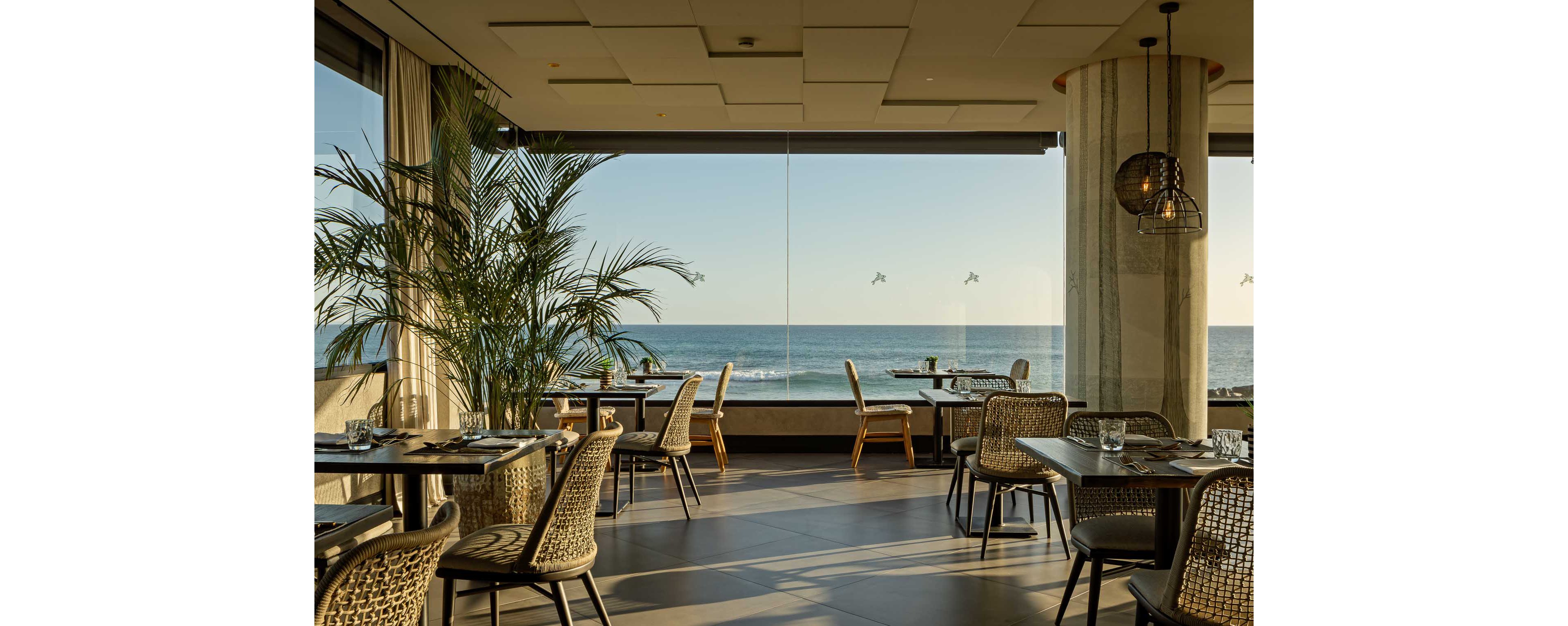 A contemporary restaurant dining area overlooks a calm ocean through large floor-to-ceiling windows. Woven chairs and neatly set tables create an inviting, relaxed atmosphere in the warm afternoon light. A tall indoor plant adds greenery, while minimalist decor and neutral tones emphasize the coastal setting. The scene is quiet and empty, suggesting readiness for guests at sunset or dinner service.