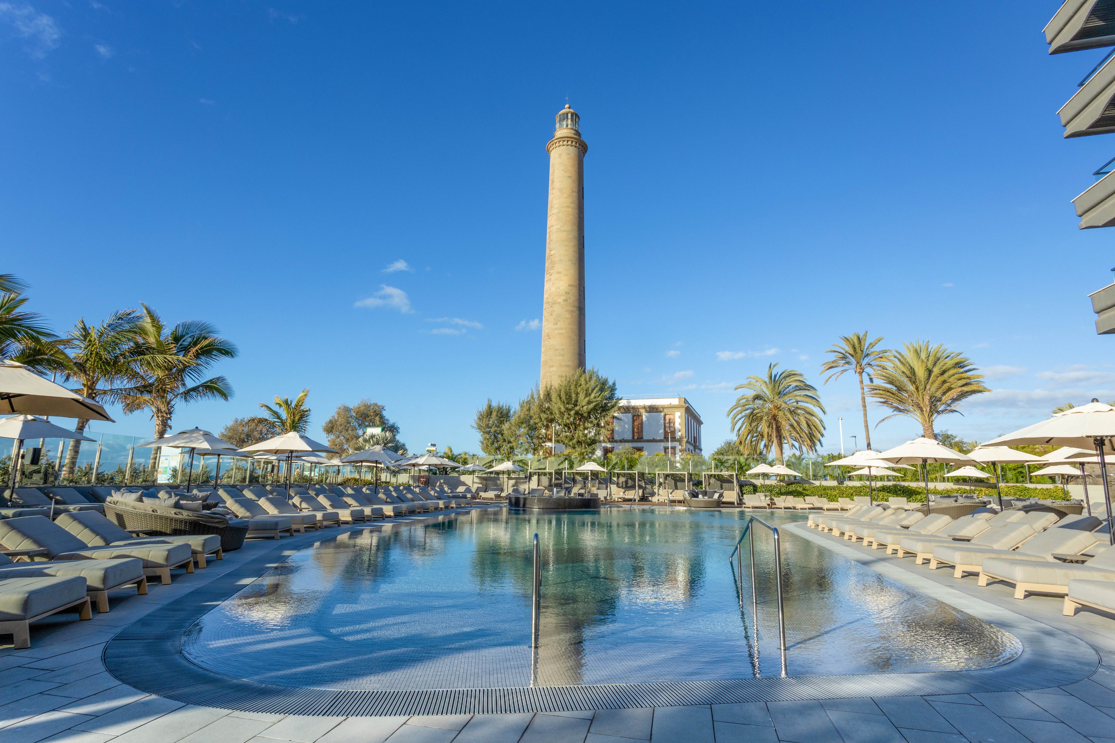 A tranquil outdoor swimming pool is surrounded by numerous lounge chairs and umbrellas, set against a backdrop of a tall lighthouse and lush palm trees. The scene is bathed in bright sunlight, with clear blue skies overhead, creating a relaxing vacation atmosphere. The pool area appears empty, emphasizing a peaceful mood and inviting setting.