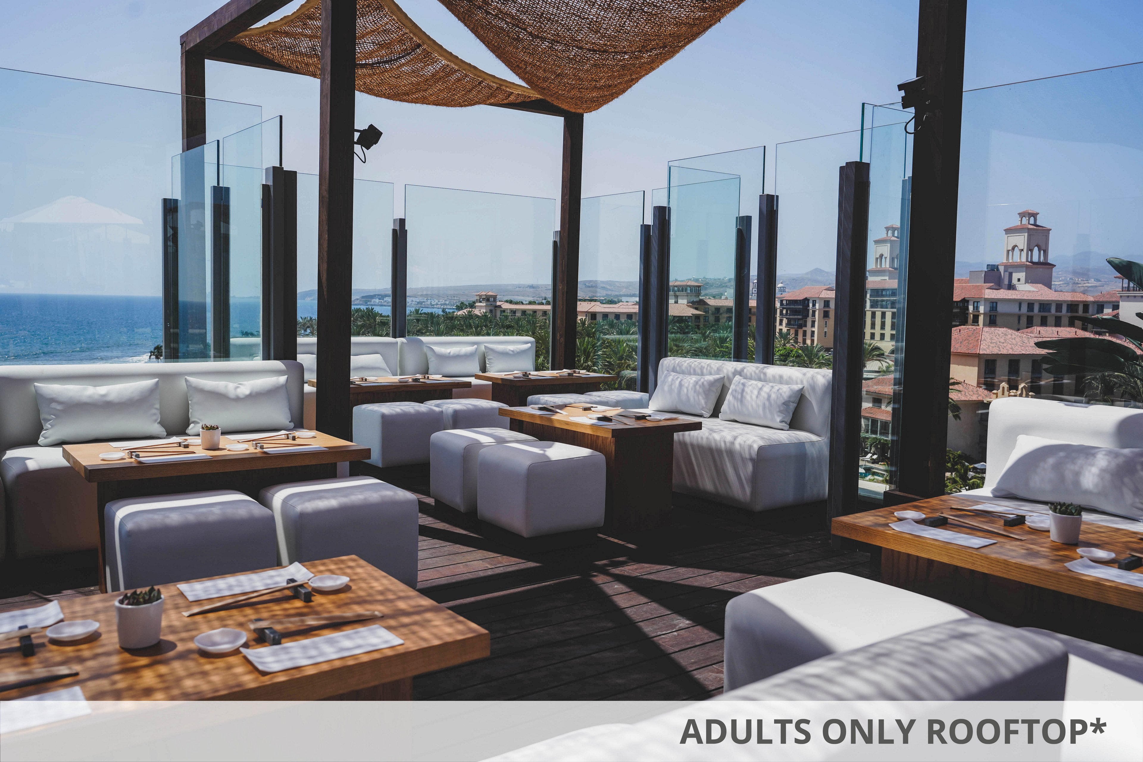 The image features a stylish rooftop lounge with white cushioned sofas and wooden tables arranged for dining. Glass panels surround the area, offering unobstructed views of the ocean and nearby buildings under a clear blue sky. The setting is bright and airy, with a contemporary design and a relaxed atmosphere. No visible text or numeric values are present in the image.