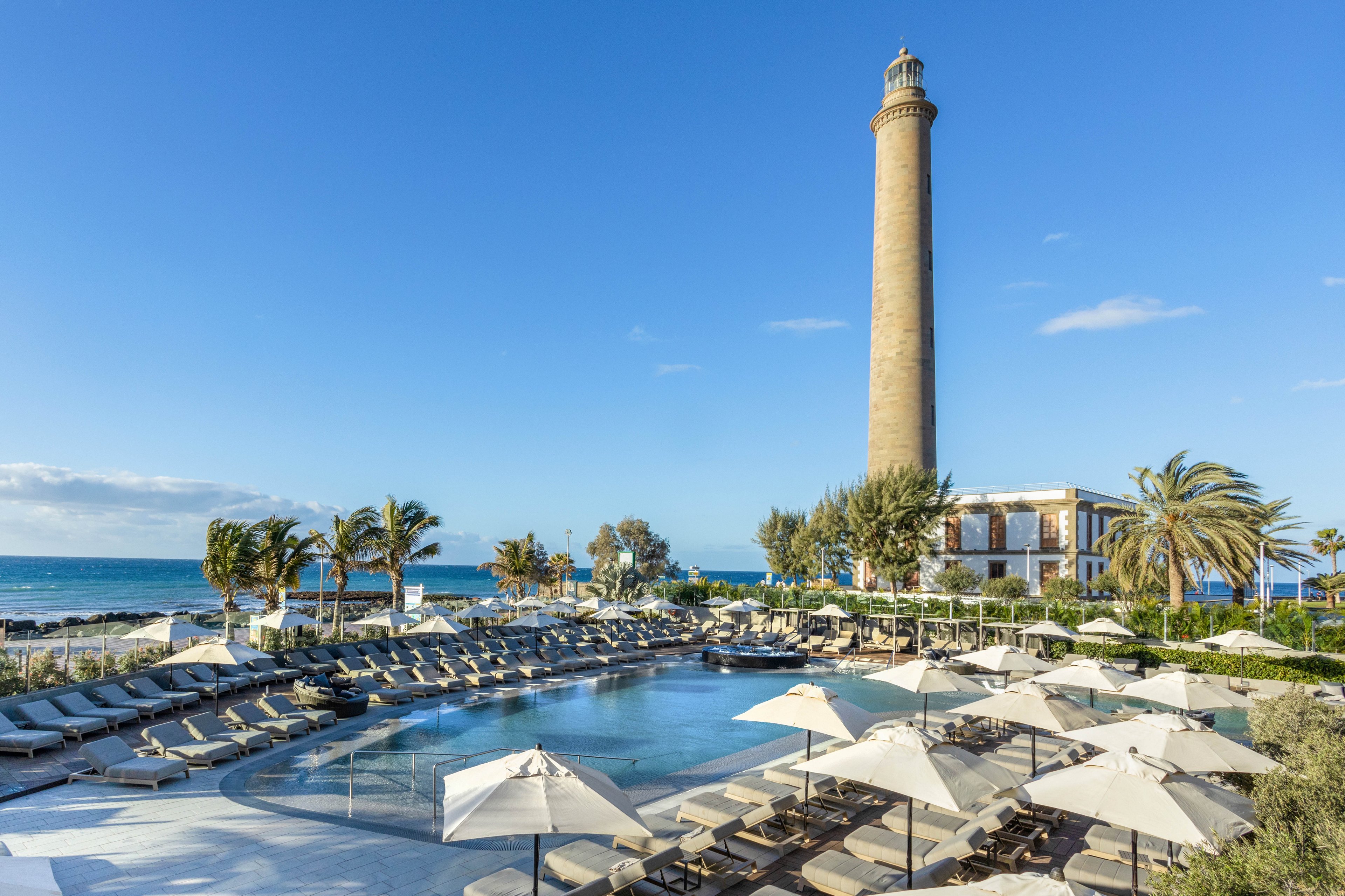 A luxurious resort pool area is shown in the foreground, surrounded by numerous lounge chairs and white umbrellas. In the background, a tall, cylindrical lighthouse stands prominently near the coastline, with the ocean and palm trees completing the scenic view. The setting is bright and tranquil, evoking a relaxing vacation atmosphere under a clear blue sky.