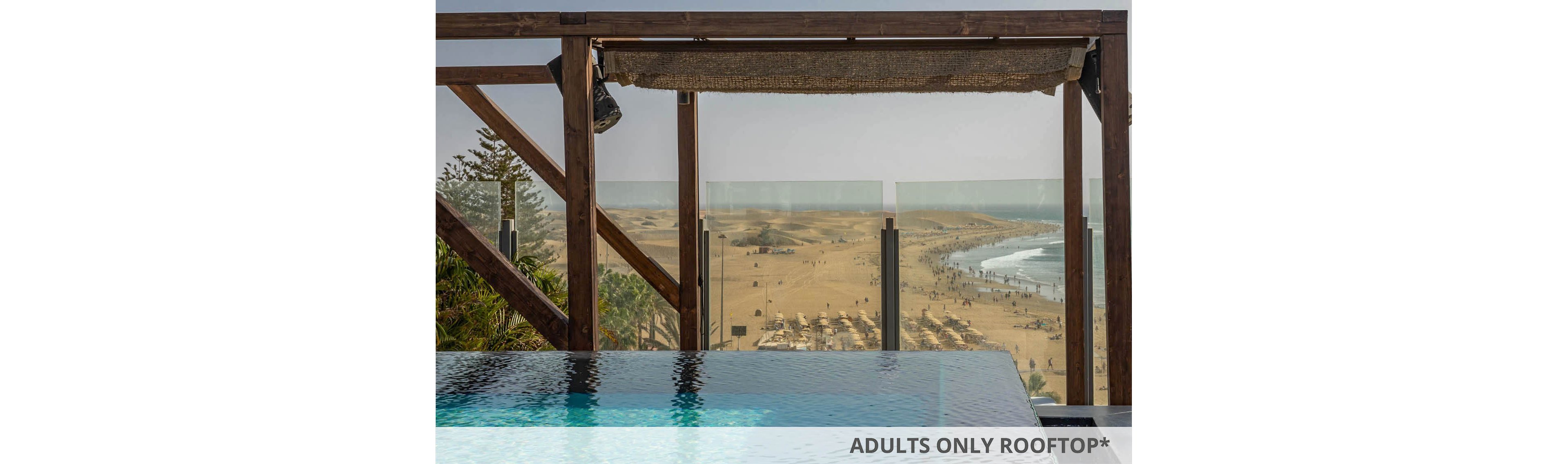 A serene infinity pool sits in the foreground, framed by a wooden pergola structure. Beyond the pool, a wide sandy beach stretches out, dotted with sun umbrellas and people enjoying the shoreline. The ocean waves gently meet the coast under a clear, bright sky, creating a tranquil and inviting atmosphere.