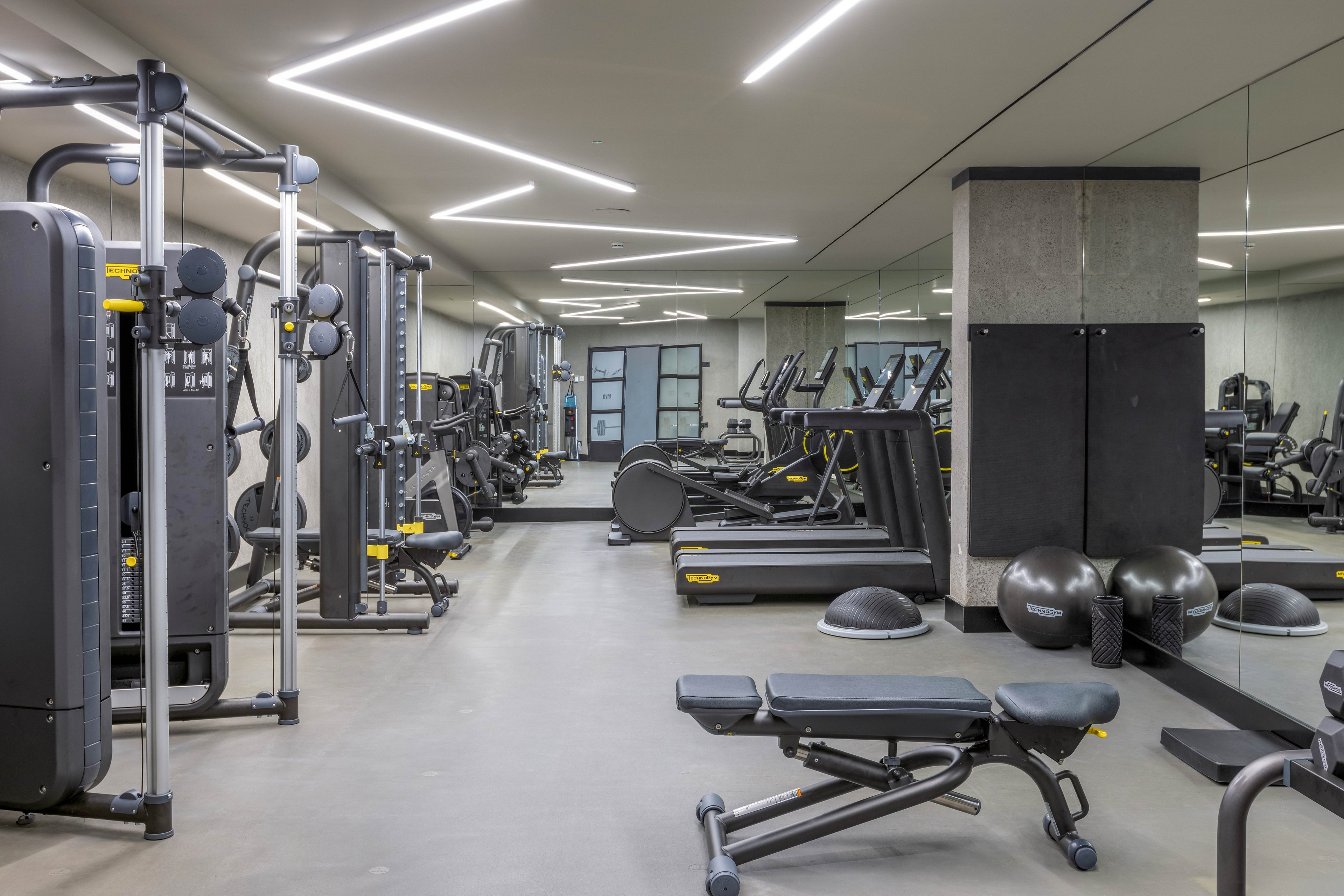 A spacious gym interior features various exercise machines, benches, and fitness accessories. The setting is clean and contemporary, with mirrored walls and geometric ceiling lights creating a bright atmosphere. Multiple treadmills, weight machines, and stability balls are visible, emphasizing a focus on strength and cardio training. No people are present, highlighting the equipment and layout.