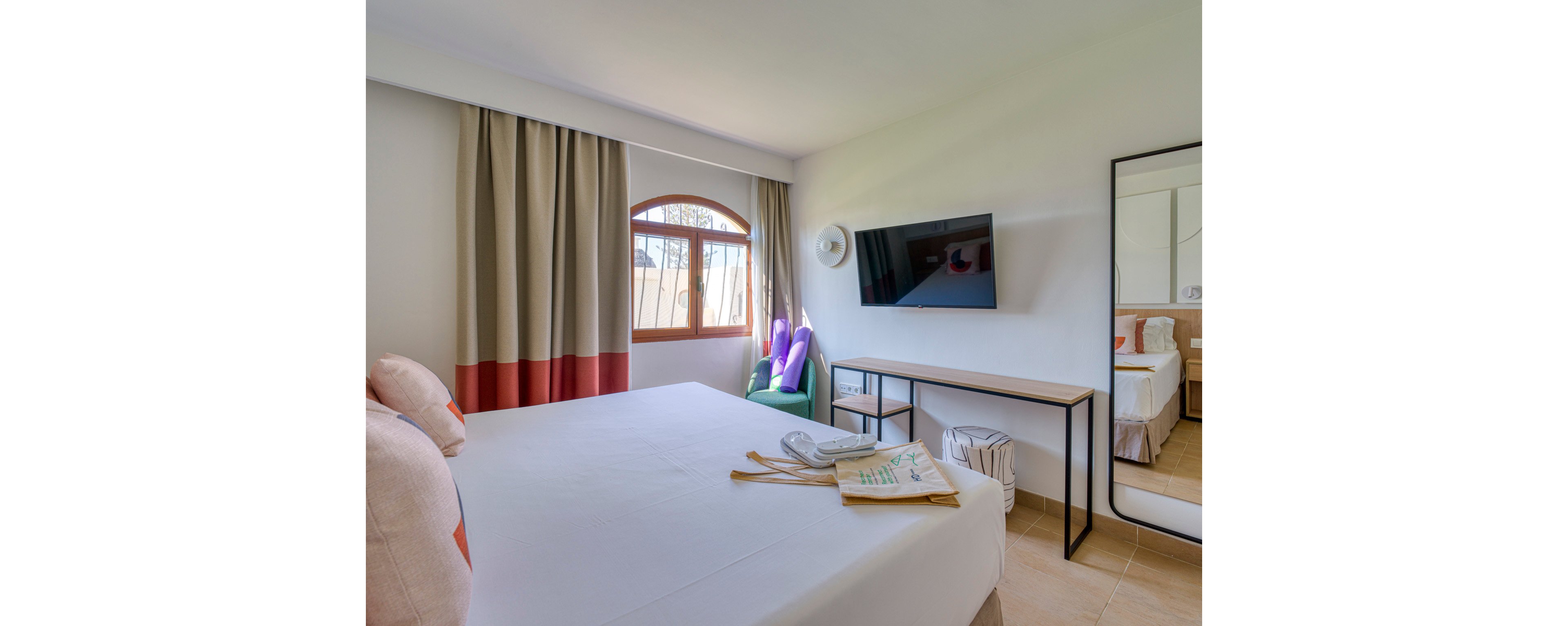 A well-lit hotel room featuring a neatly made bed, a wall-mounted flat-screen TV, and a desk with a mirror. The room includes colorful curtains, a green armchair, and a window allowing natural light. A telephone and a booklet are placed on the bed, adding to the cozy ambiance.