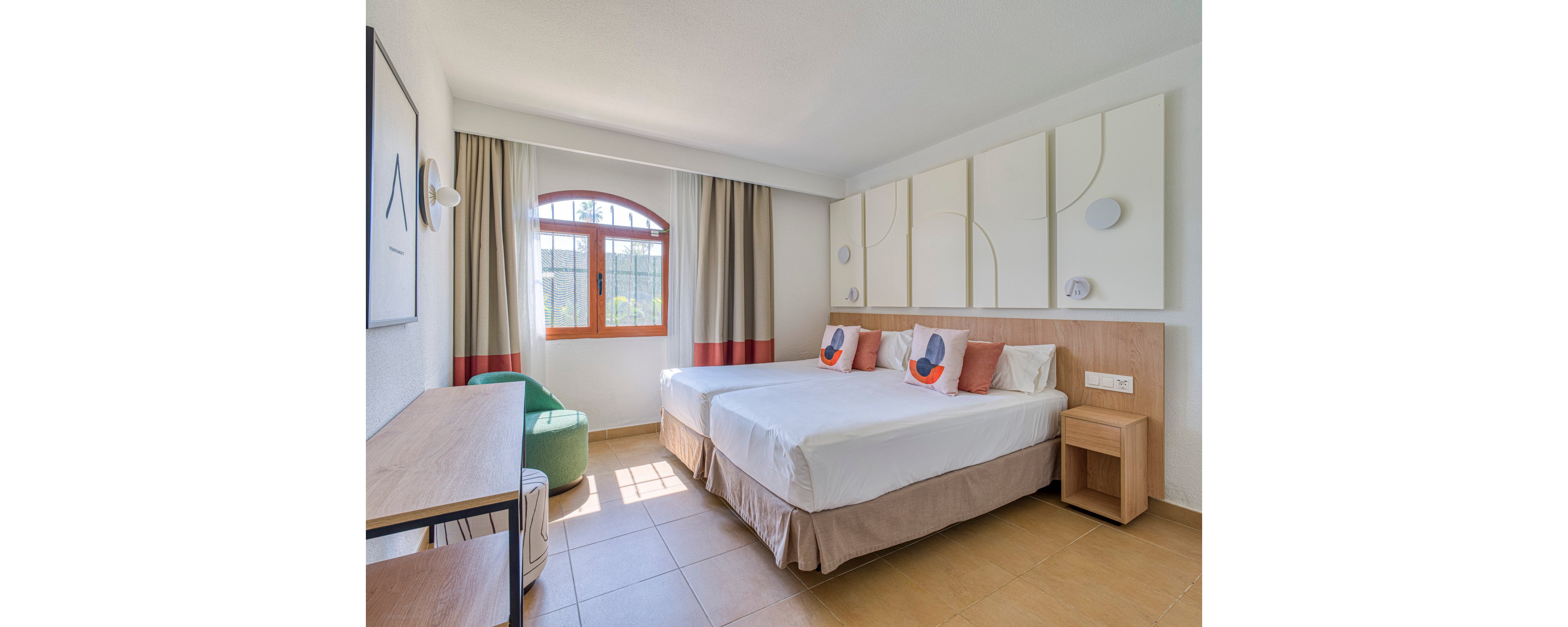 A bright, contemporary hotel room features two neatly made twin beds with white linens and colorful accent pillows. Natural light streams through a wooden-framed window with beige and red curtains, illuminating the tiled floor. Minimalist wall art and paneling decorate the headboard area, while a small bedside table and desk provide functional storage. A green upholstered chair adds a pop of color, creating a clean and inviting atmosphere.