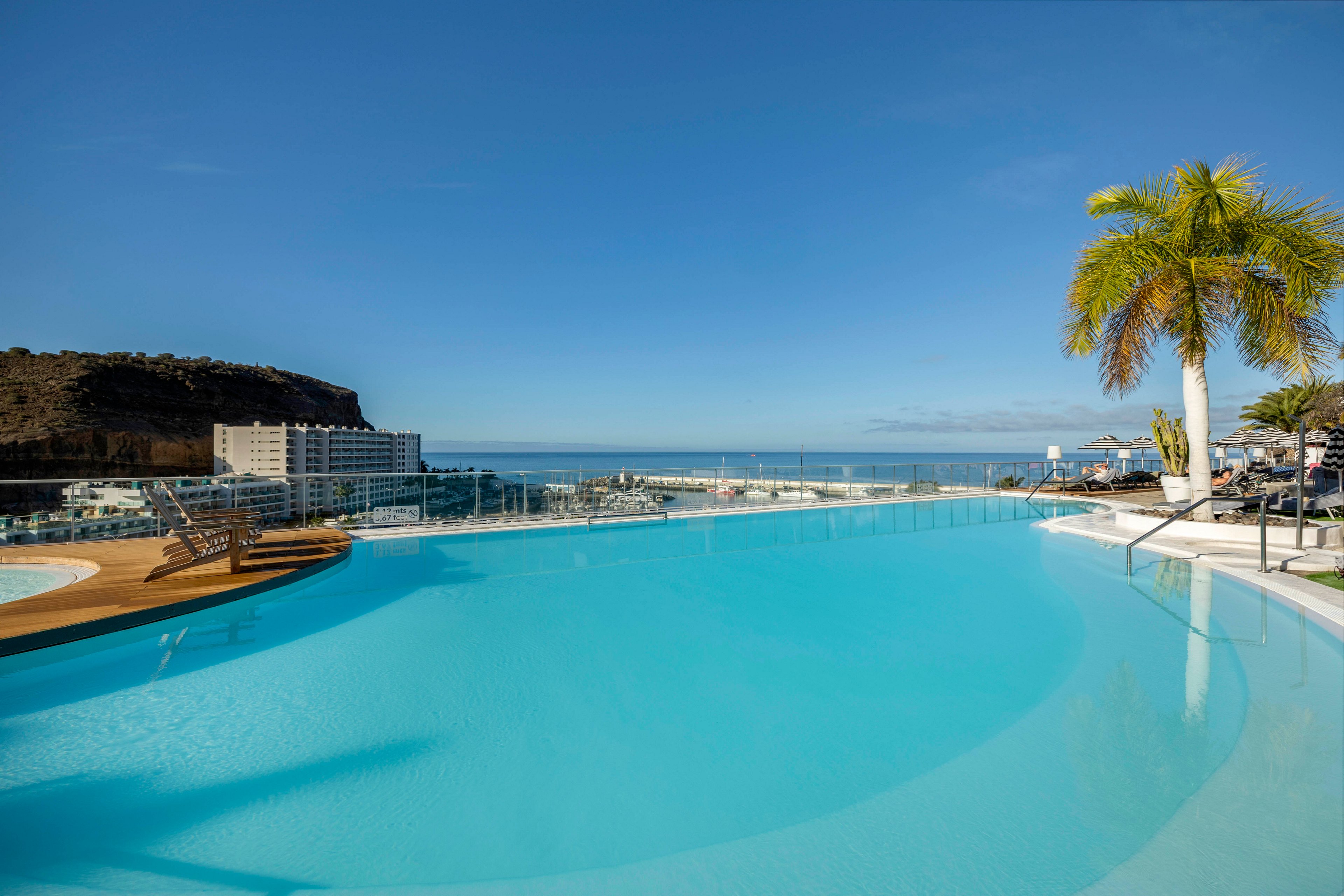 A large infinity pool stretches across the foreground, reflecting the clear blue sky. Sun loungers and a wooden deck line the poolside area, suggesting a relaxing resort setting. In the distance, modern buildings and a rocky hillside rise above the coastline. The calm ocean and palm trees create a tranquil, tropical atmosphere under bright daylight.