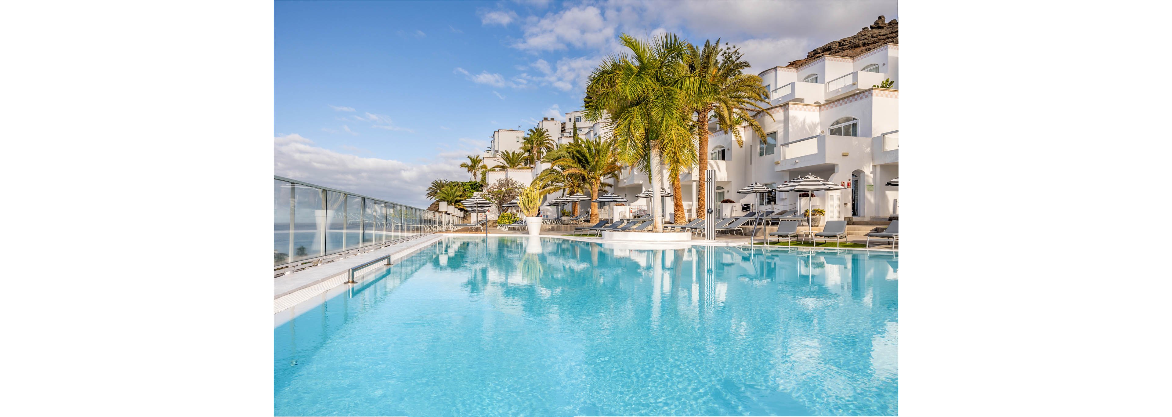 A large turquoise swimming pool stretches alongside a modern seaside resort with white buildings. Tall palm trees and sun loungers line the pool deck, creating a relaxing vacation atmosphere. Glass railings reveal an ocean view under a bright blue sky with scattered clouds. The scene suggests a tranquil coastal getaway with warm, sunny weather.