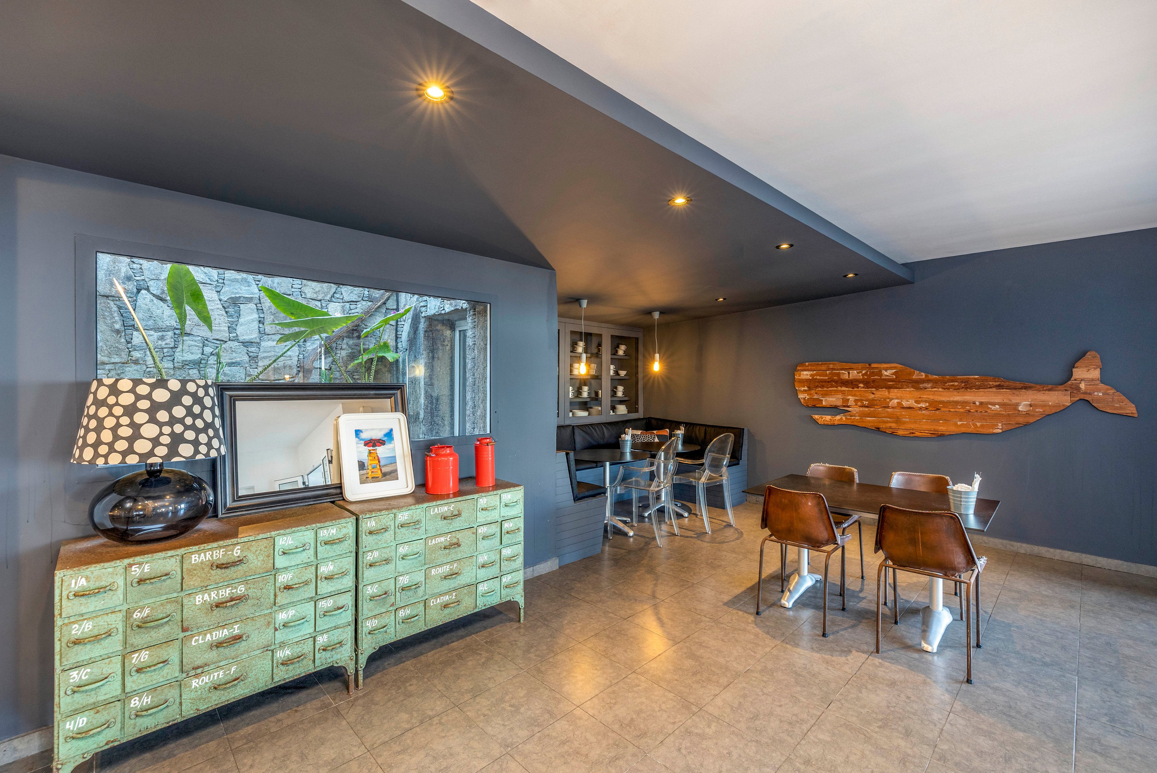 A contemporary dining room features a small table with four brown leather chairs set against a dark gray wall. On the left, a mint green sideboard holds a patterned table lamp, framed artwork, and two red decorative containers beneath a large window. In the background, a cozy nook with a bar-height table and metal chairs sits under recessed ceiling lights. A large wooden whale wall art piece adds a coastal touch to the minimalist interior. The overall palette combines cool grays with warm wood tones and colorful accents.