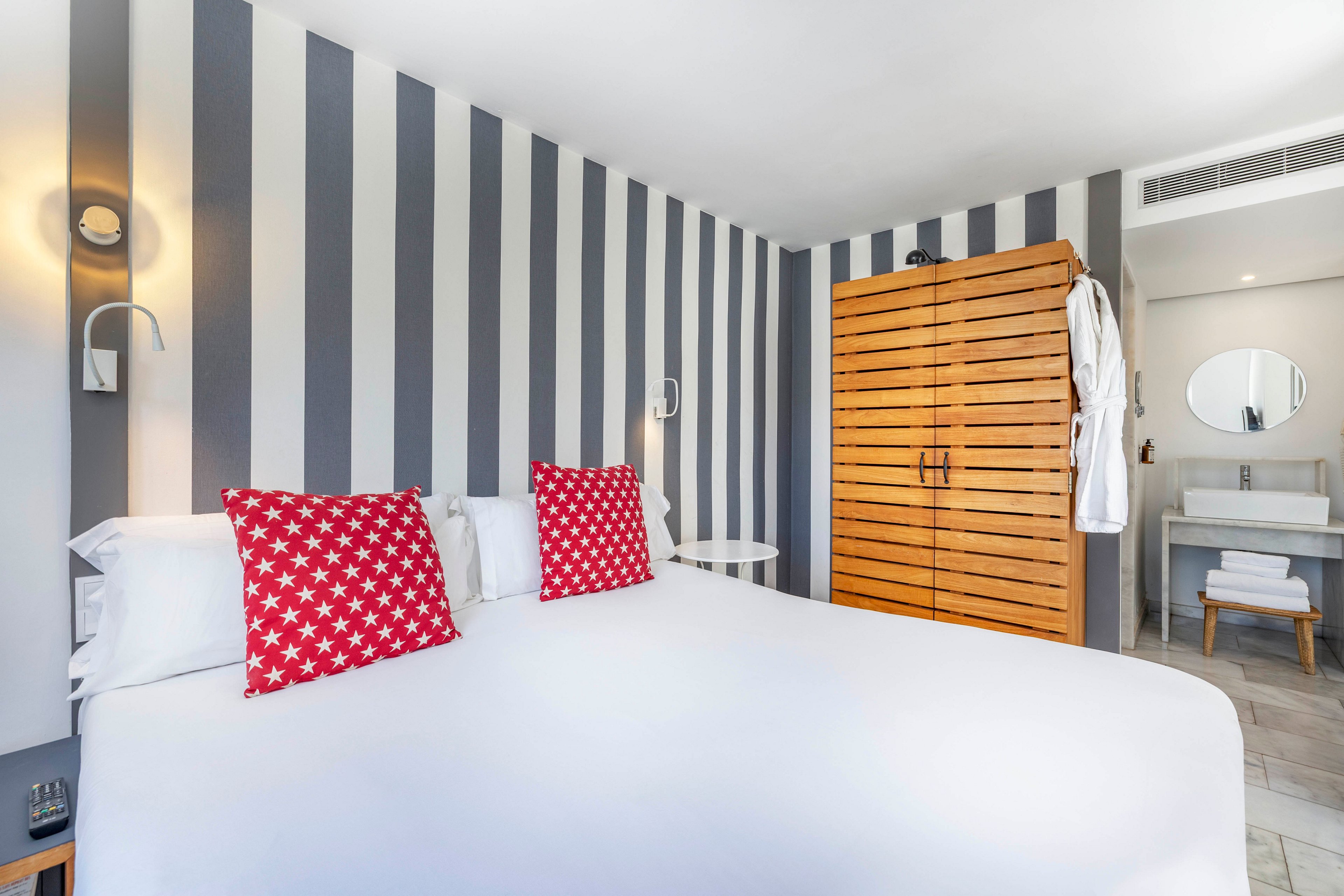 A bright modern hotel bedroom features a large neatly made bed with crisp white linens and red star-patterned pillows. The focal point is a bold vertical striped accent wall in shades of gray and white behind the bed. To the side, a wooden slatted wardrobe and hanging white bathrobes add warmth and texture. An open doorway reveals a minimalist bathroom with a round mirror and folded towels, enhancing the clean, contemporary feel of the space.