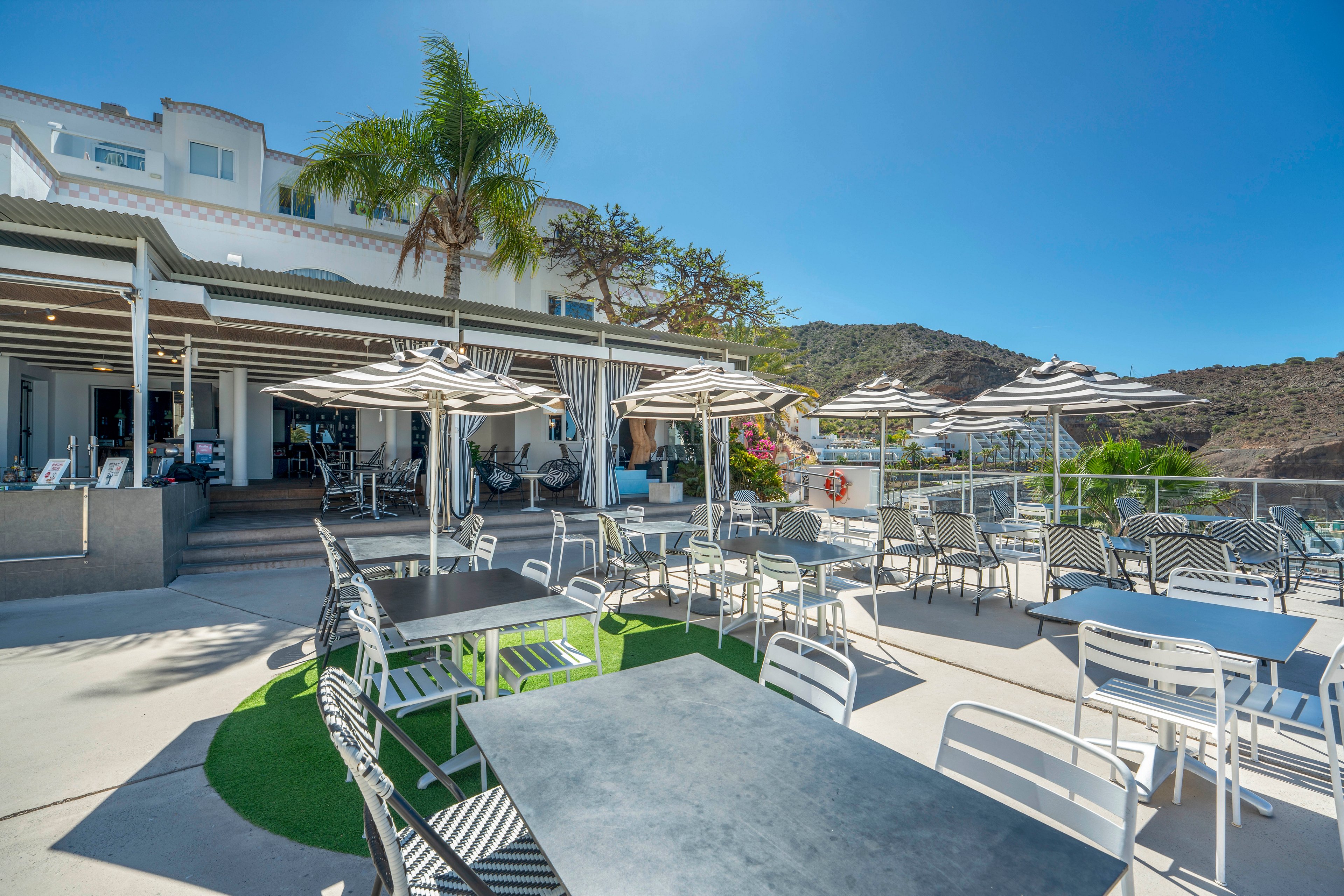 An outdoor cafe terrace features metal tables and chairs arranged under striped umbrellas. The setting is a bright, sunny day with clear blue skies and a modern white building in the background. Palm trees and surrounding hills create a relaxed, resort-like atmosphere. The scene appears quiet and inviting, ideal for outdoor dining or lounging by the patio.