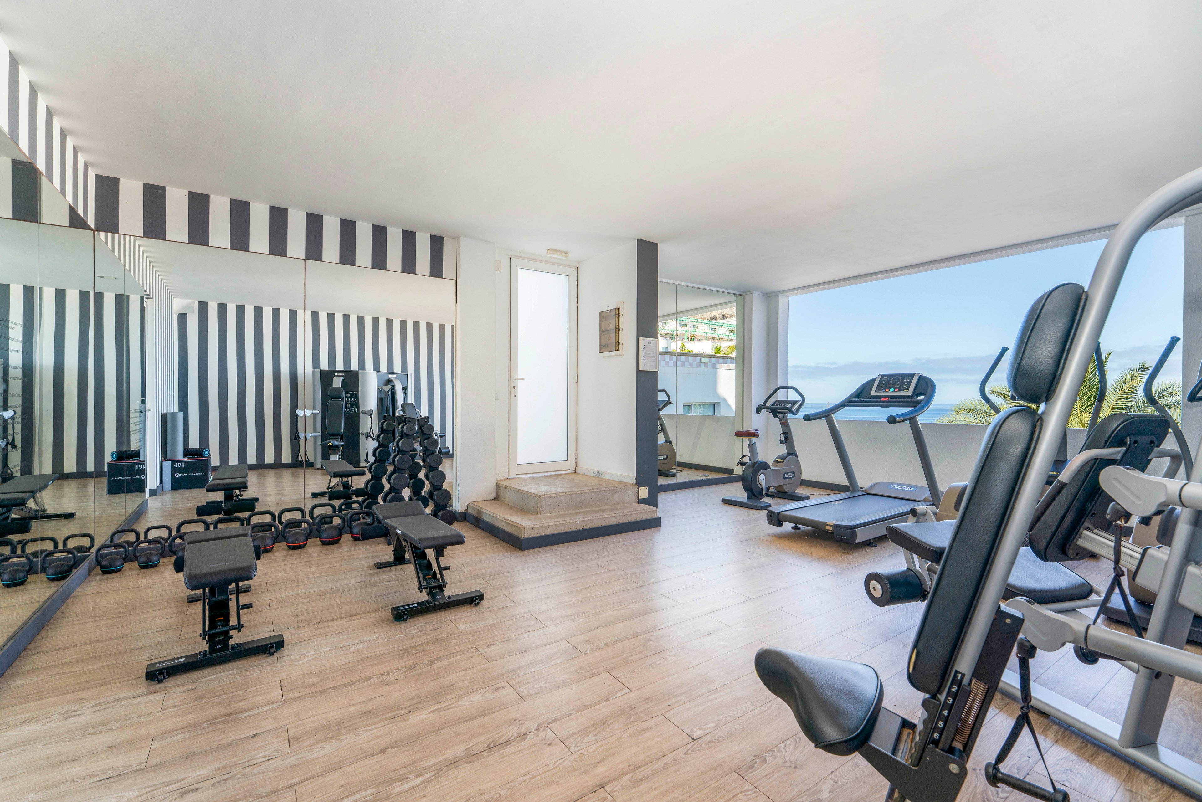 A bright, modern fitness room features various exercise machines and free weights. Cardio equipment such as treadmills and stationary bikes face large windows with a scenic ocean view. Benches and dumbbells are arranged neatly on a light wooden floor, with mirrors and striped wall panels enhancing the spacious feel. The clean, minimalist design suggests a contemporary hotel or apartment gym focused on wellness and relaxation.