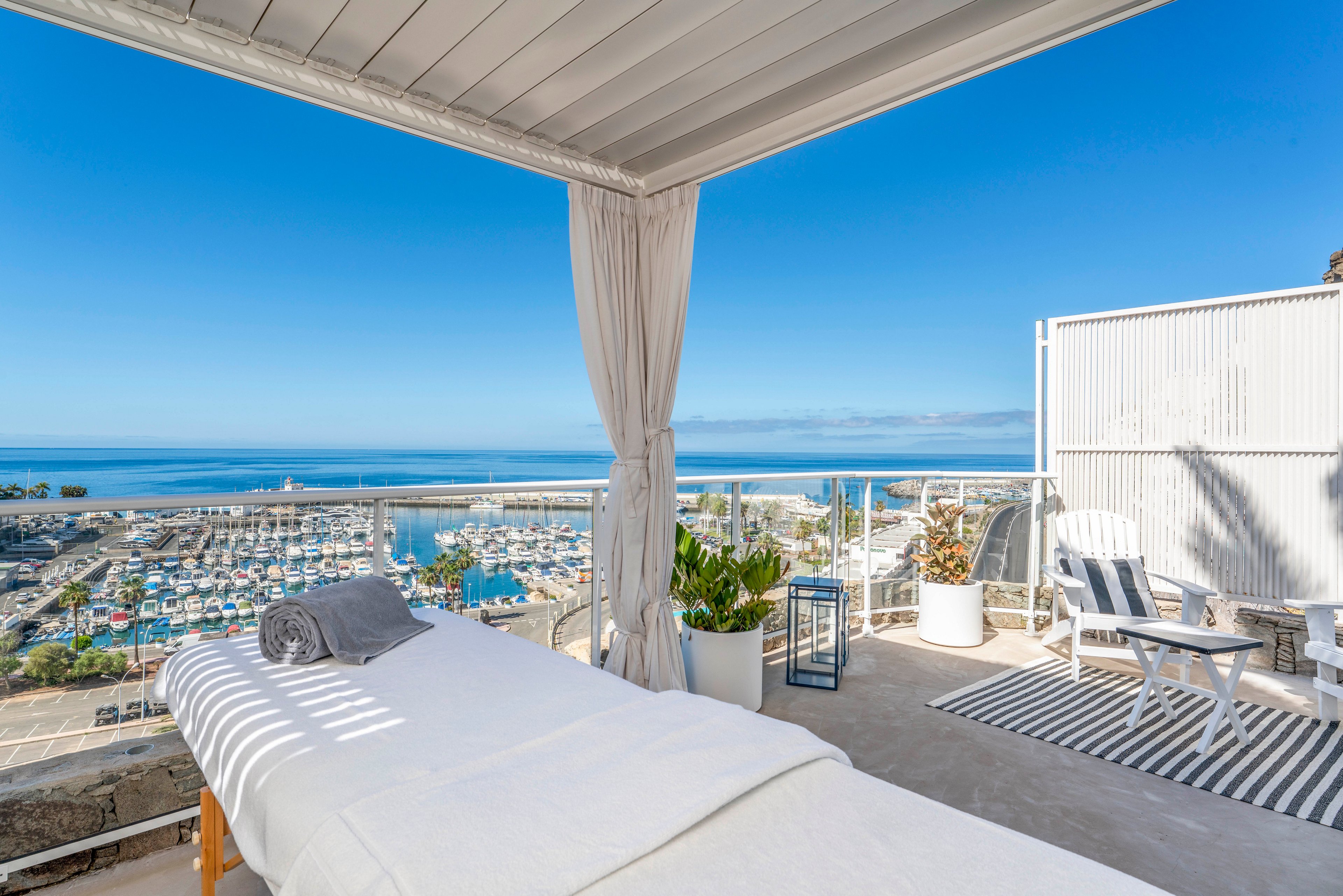 A massage bed is set up in a covered outdoor cabana with a view of a marina and the open sea. The bright, sunny setting features clear blue skies and calm water in the distance. Potted plants, a striped rug, and white lounge chairs add a relaxed, coastal decor. The scene evokes a tranquil spa experience in a luxurious seaside location.
