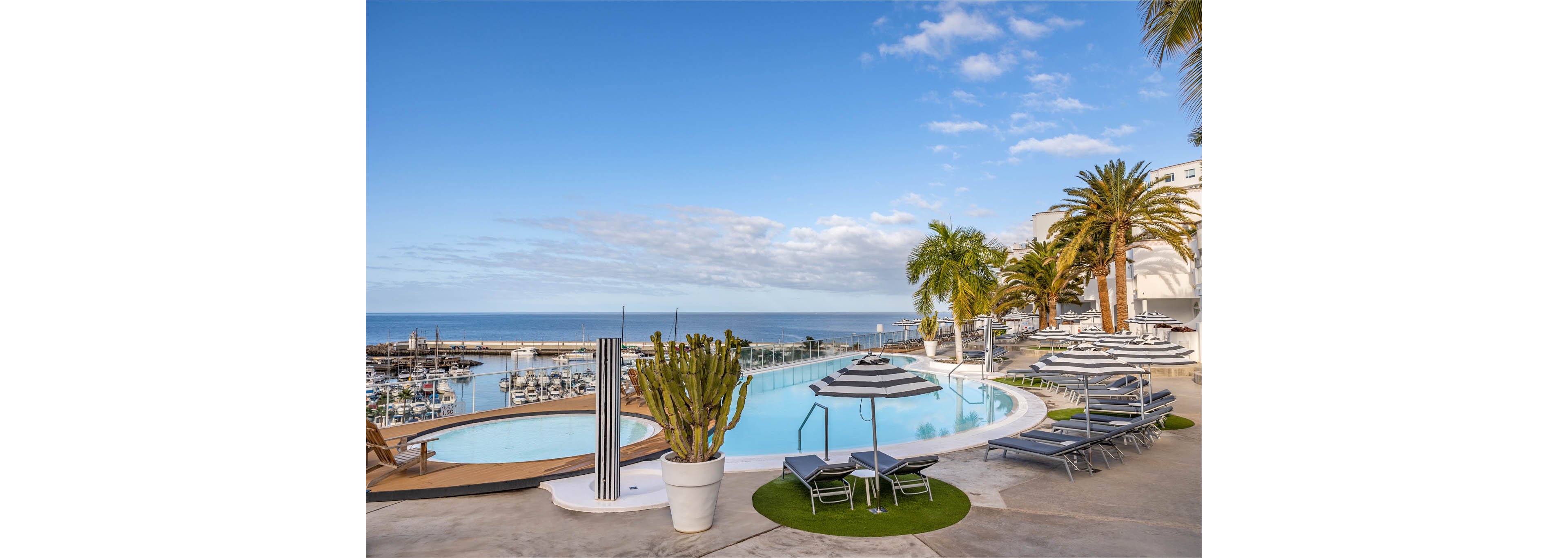 A contemporary resort swimming pool curves along a terrace above a calm marina. Sun loungers and parasols line the poolside, facing the ocean horizon under a bright blue sky. Palm trees and a large potted cactus add tropical greenery to the sleek white architecture. The scene feels tranquil and luxurious, ideal for a coastal vacation or waterfront relaxation.
