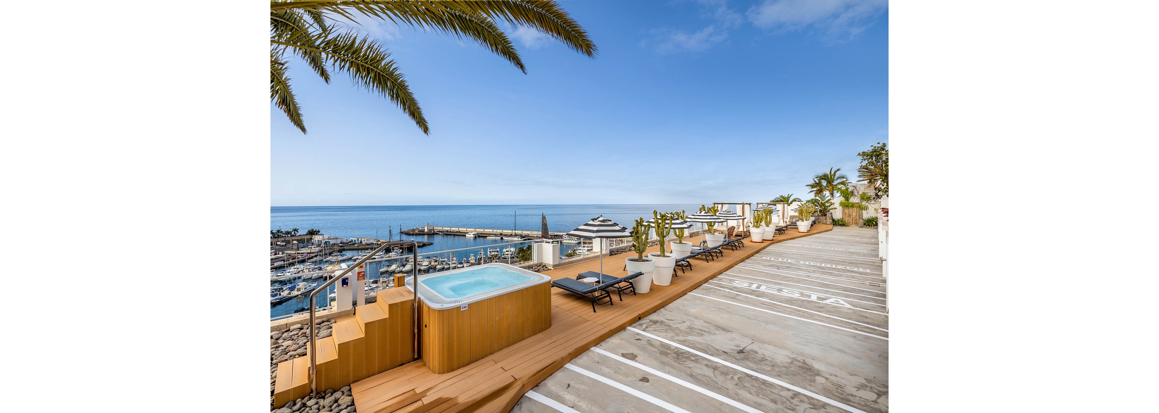 A rooftop terrace features a wooden deck with a hot tub and several lounge chairs facing the sea. The setting overlooks a marina filled with boats and a distant breakwater under a clear blue sky. Potted palm trees and cacti line the deck, adding a tropical feel to the modern outdoor space. An adjacent concrete area with painted white lines suggests parking or multipurpose use near the terrace.