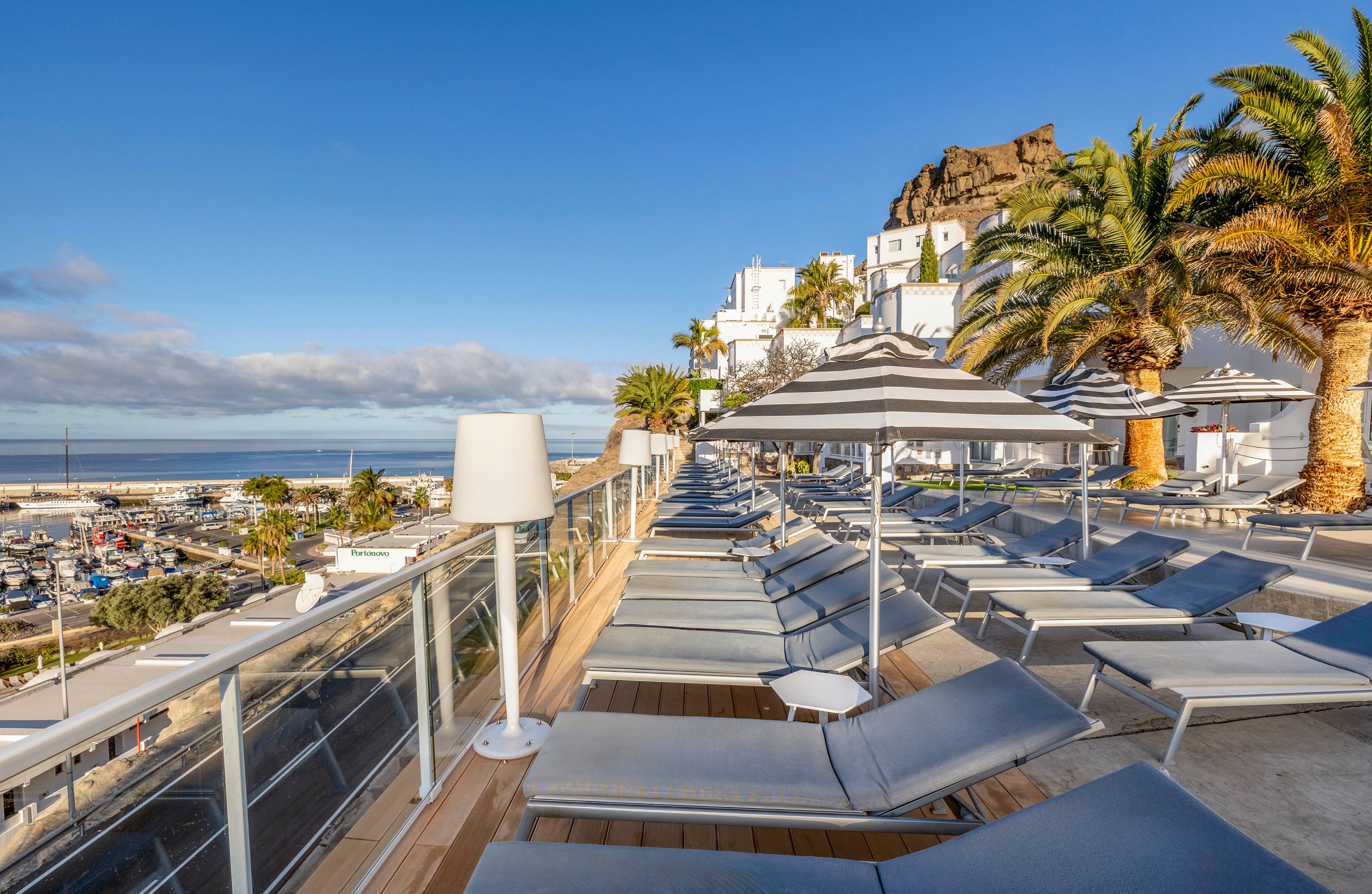 A modern seaside resort terrace is lined with empty sun loungers facing the ocean. Striped parasols and contemporary lamps create a stylish, orderly arrangement along the deck. Palm trees and white hillside buildings frame the scene, suggesting a Mediterranean or coastal holiday destination. The clear blue sky and calm water enhance the relaxed, vacation atmosphere.
