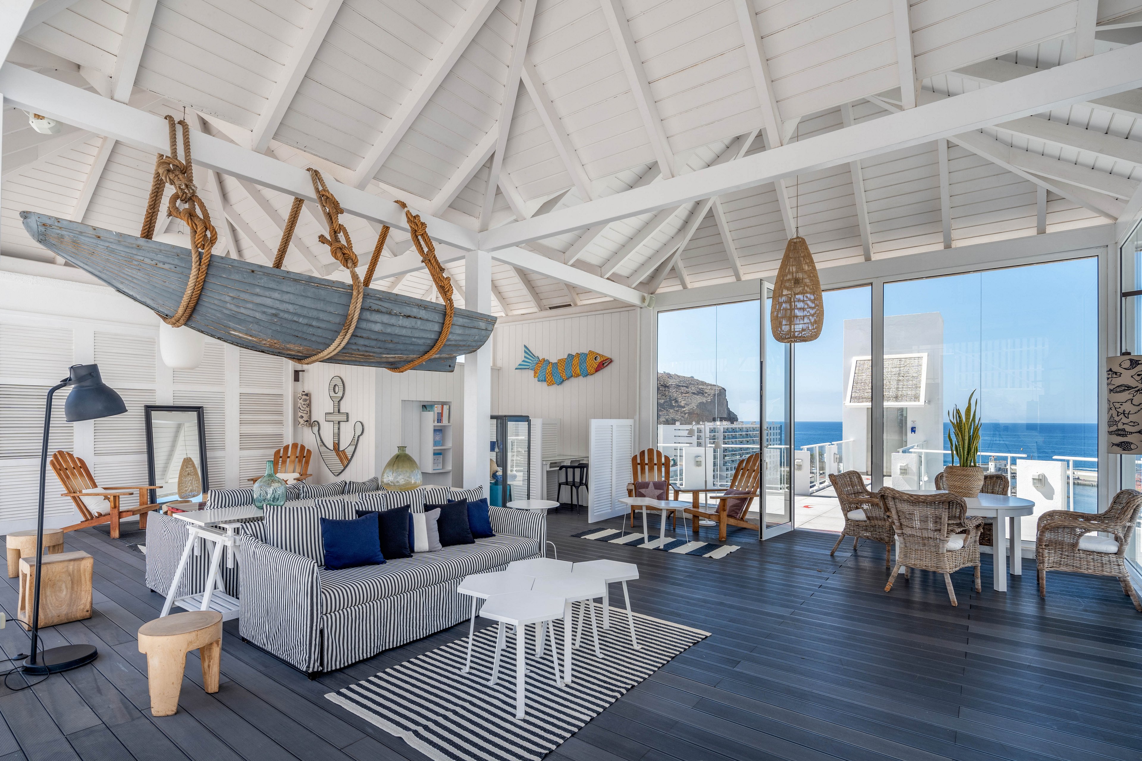 Bright coastal-style living room featuring a striped sofa and nautical decor. A suspended wooden boat and anchor artwork emphasize the seaside theme. Large windows reveal a clear ocean view and rocky coastline outside. Wicker chairs, wooden stools, and striped rugs add texture and warmth to the airy white space.
