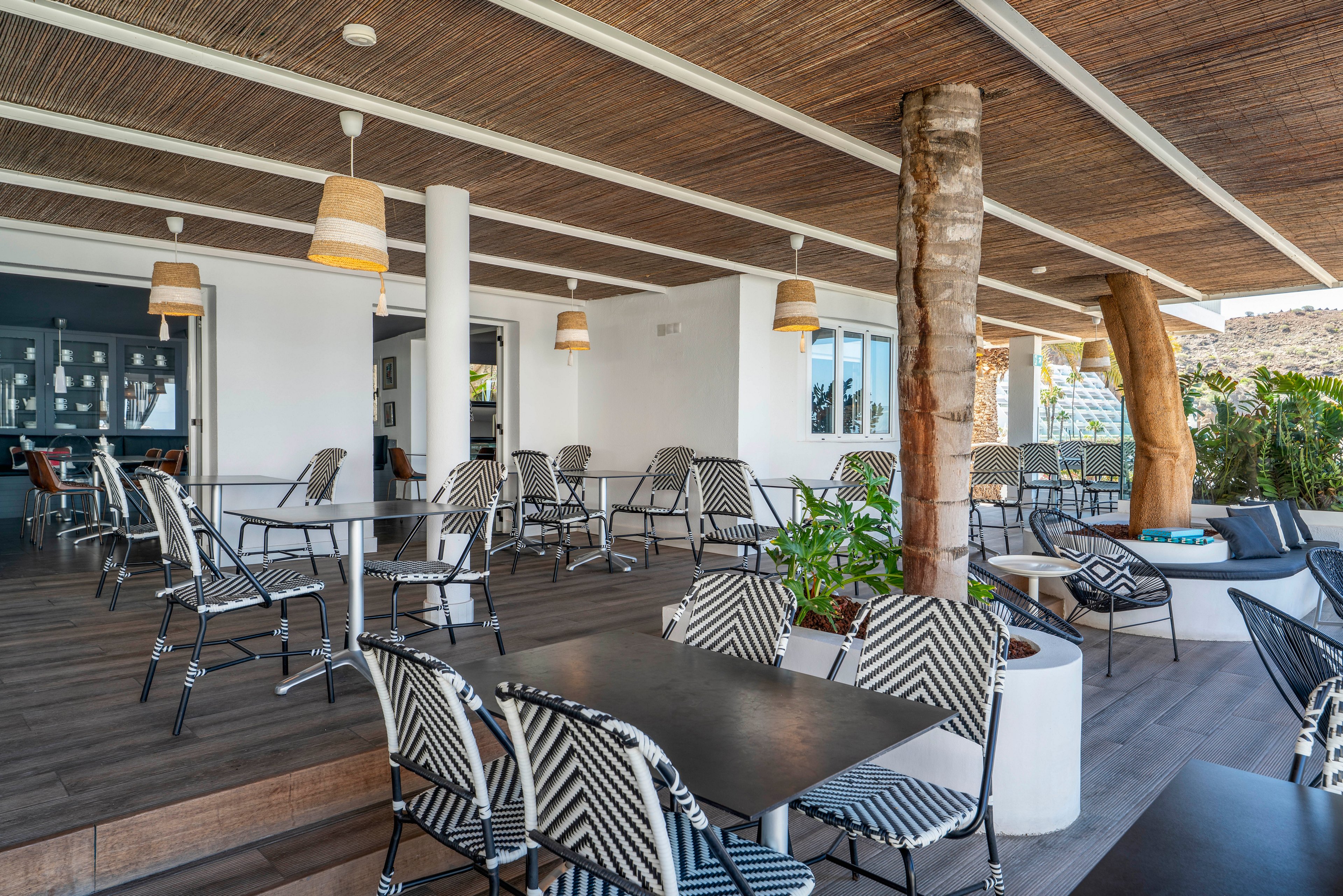 An open-air terrace café features neatly arranged black tables and patterned chairs. The space is covered by a wooden ceiling with hanging woven lamps, creating a shaded and inviting atmosphere. Large white columns and tree trunks integrate natural elements into the modern design. In the background, glass doors and greenery suggest a relaxed coastal or resort setting.