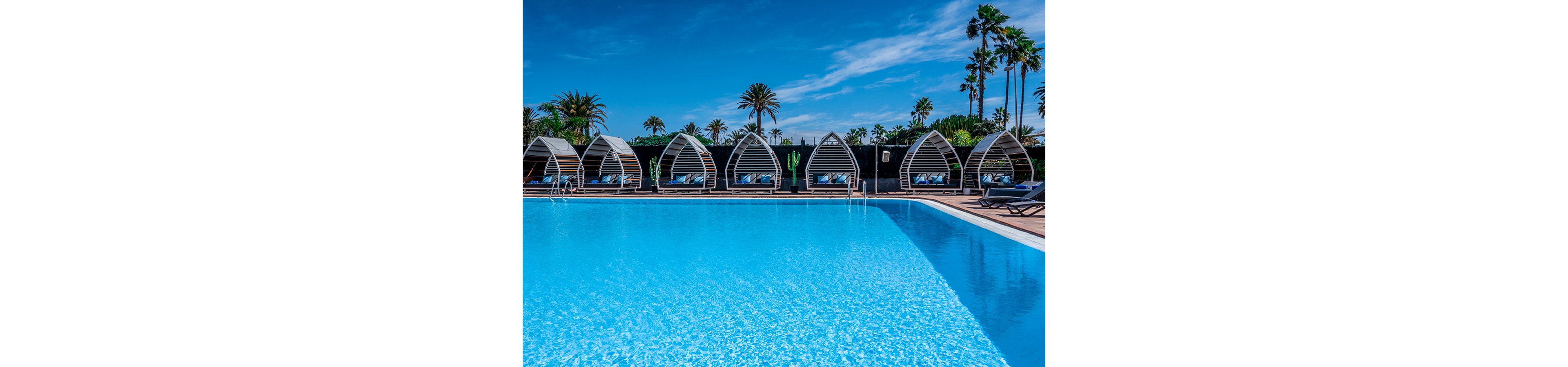 A bright blue swimming pool dominates the foreground of a stylish resort scene. Behind the pool, a row of contemporary white cabanas with curved frames and lounge chairs sit neatly aligned. Tall palm trees rise above a dark fence, contrasting with the vivid blue sky and scattered clouds. The overall atmosphere is sunny, relaxing, and ideal for a tropical vacation.