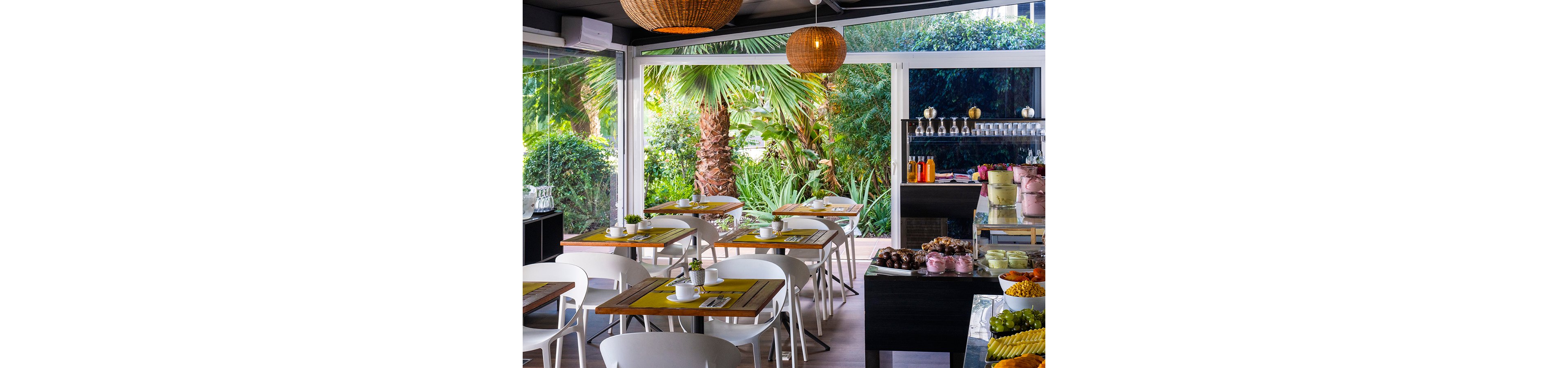 A modern café dining area features wooden tables with white chairs and yellow placemats. Large glass windows open onto lush tropical greenery with palm trees, creating a vibrant garden backdrop. A buffet counter displays assorted desserts, pastries, fruits, and beverages in colorful bowls and jars. Warm hanging wicker lamps add a cozy, inviting atmosphere to the bright interior.