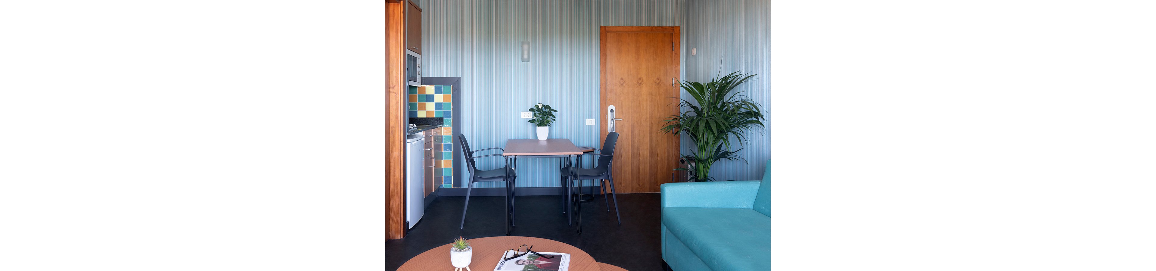 Compact contemporary living space featuring a wooden dining table with two black chairs. A turquoise sofa sits in the foreground beside a large potted plant, while a small kitchenette with colorful tiled backsplash is visible on the left. The room has light blue striped walls, a wooden door, and several potted plants adding a fresh touch. A round coffee table holds a magazine and eyeglasses, contributing to a relaxed, homey atmosphere.