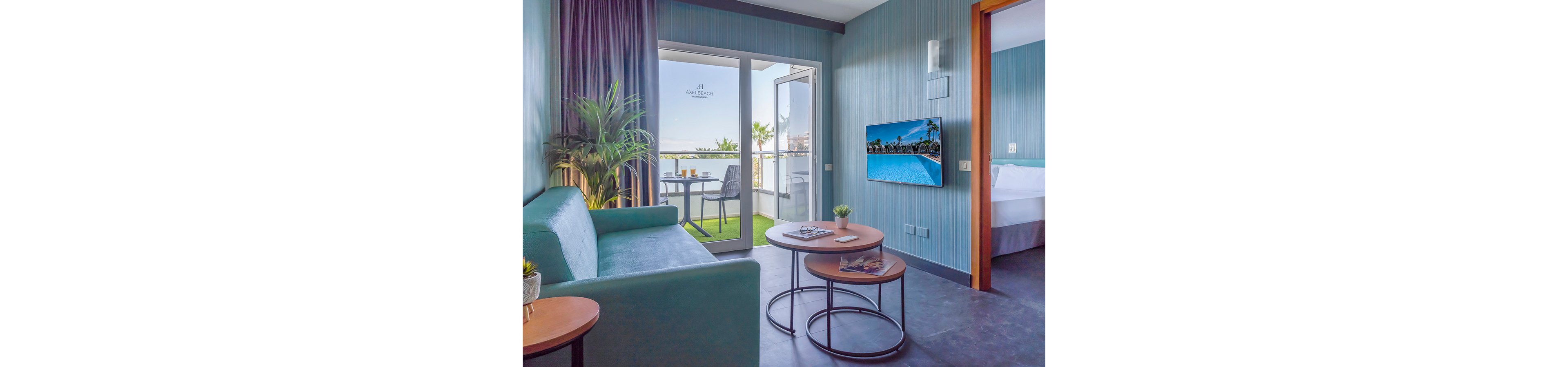 Contemporary hotel suite features a teal sofa and nested round coffee tables in a bright living area. A wall-mounted television displays a swimming pool scene, adding a leisure atmosphere to the room. Glass doors open onto a balcony with outdoor seating and potted plants, overlooking a coastal view. The adjoining bedroom with a neatly made bed is partially visible through the open doorway, enhancing the sense of space and comfort.