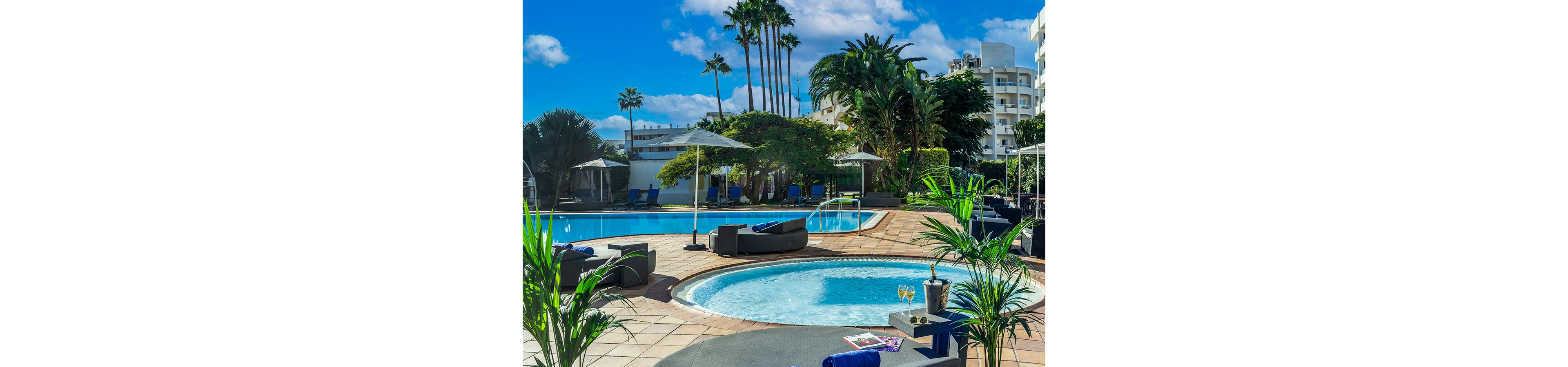 Outdoor resort pool area featuring a round jacuzzi in the foreground and a large swimming pool behind it. Modern lounge chairs and umbrellas are arranged on the tiled terrace, with tropical palm trees and lush greenery surrounding the space. High-rise buildings frame the background under a bright blue sky with scattered clouds. A small table holds two glasses of white wine, an ice bucket, and magazines, adding to the relaxed vacation atmosphere.