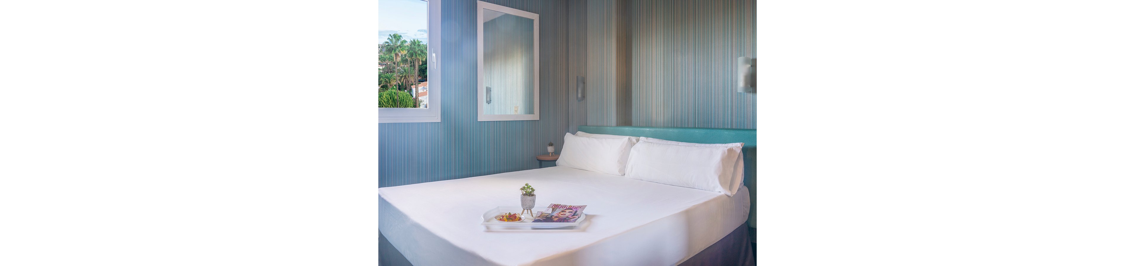 A neatly made double bed with white linens sits in a modern, light-filled bedroom. Striped blue wallpaper and a large window with a view of greenery create a fresh, contemporary atmosphere. A bedside table and wall-mounted lights add functional comfort, while a tray with magazines and a small potted plant rests on the bed. The overall scene suggests a clean, inviting hotel or guest room ready for relaxation.