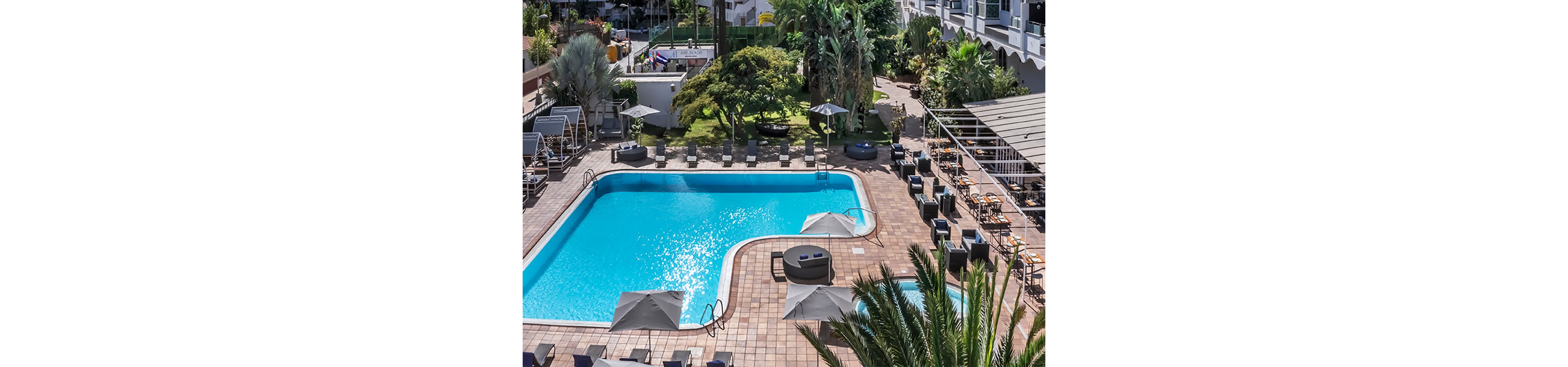 An outdoor hotel swimming pool is surrounded by lounge chairs and umbrellas in a tiled courtyard. Tall palm trees and lush greenery frame the pool area, adding a tropical atmosphere. On one side, a bar or seating area with tables and stools is arranged under a canopy. The setting appears calm and inviting, suitable for vacation relaxation and leisure.