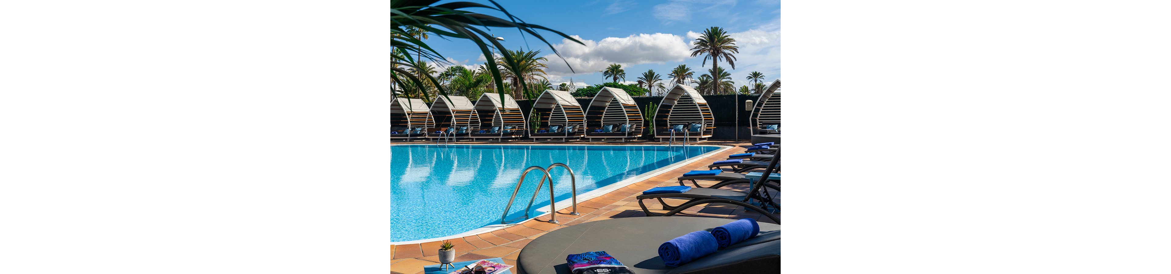 A bright outdoor swimming pool is surrounded by stylish lounge chairs and contemporary cabanas. Rolled blue towels and a small potted plant rest on a sunbed in the foreground, suggesting a relaxing vacation setting. Palm trees and lush greenery frame the pool area under a partly cloudy blue sky. The scene conveys a tranquil, upscale resort atmosphere ideal for leisure and sunbathing.