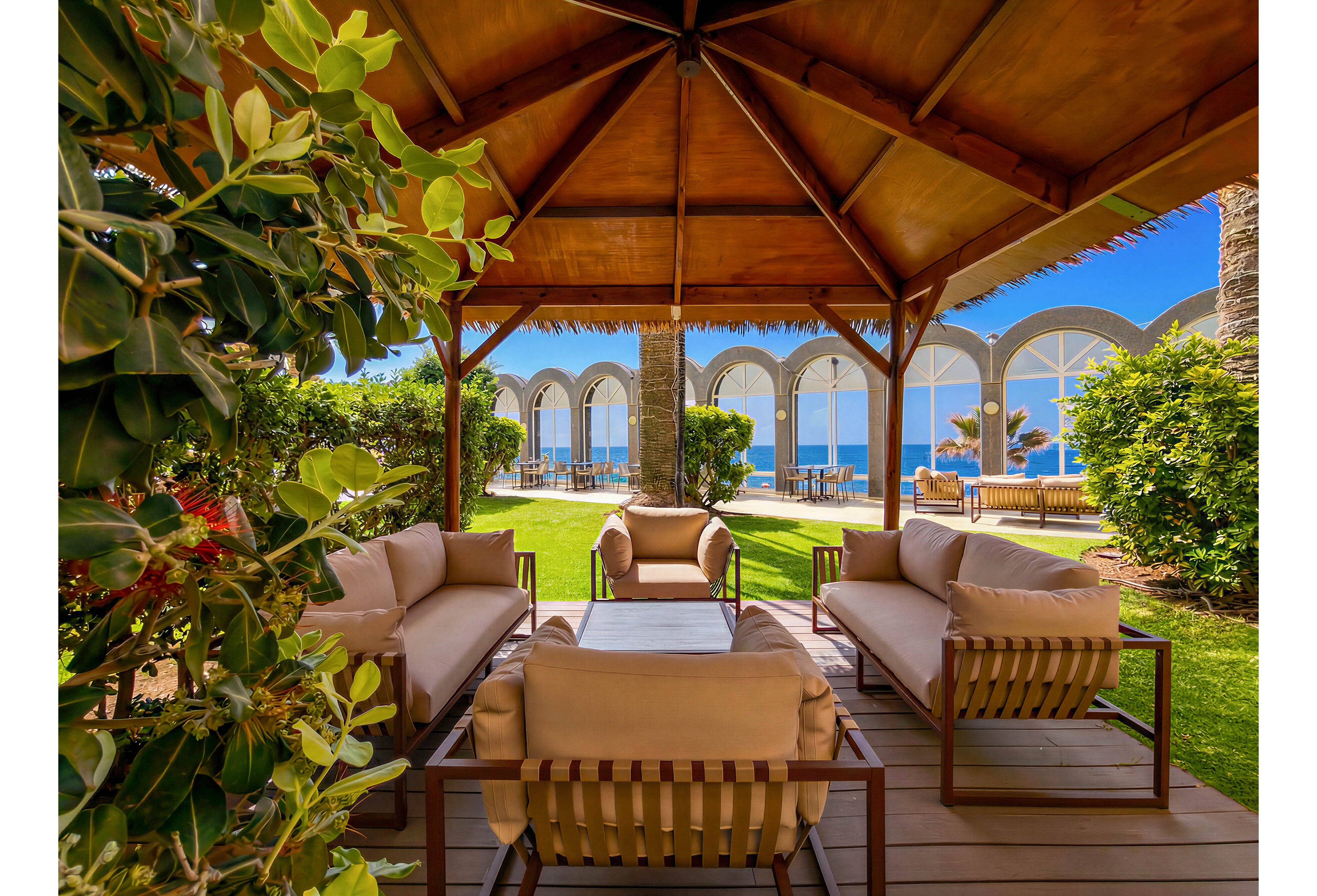 An inviting outdoor lounge area is arranged beneath a wooden gazebo with cushioned sofas and armchairs. The seating overlooks a manicured lawn leading to arched glass structures and a view of the sea. Lush green shrubs and palm trees frame the scene, adding a tropical resort atmosphere. Bright sunlight and clear blue skies create a warm, relaxing vacation mood.