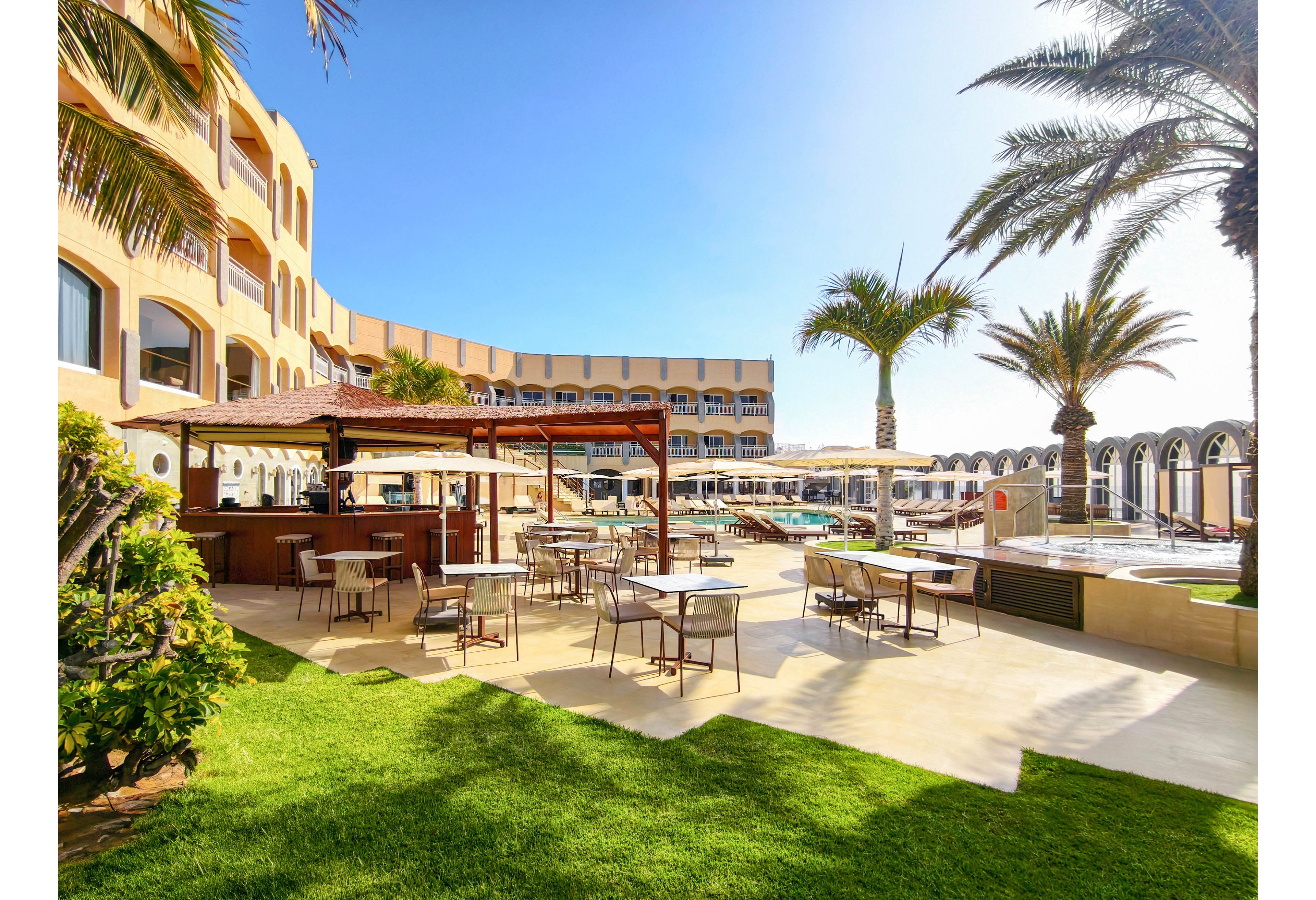 An outdoor resort terrace features tables and chairs arranged beside a wooden bar area. A large swimming pool stretches along the curved hotel building in the background. Tall palm trees and bright green grass add a tropical feel under a clear blue sky. The scene suggests a relaxed vacation atmosphere with ample seating and sunlit open spaces.