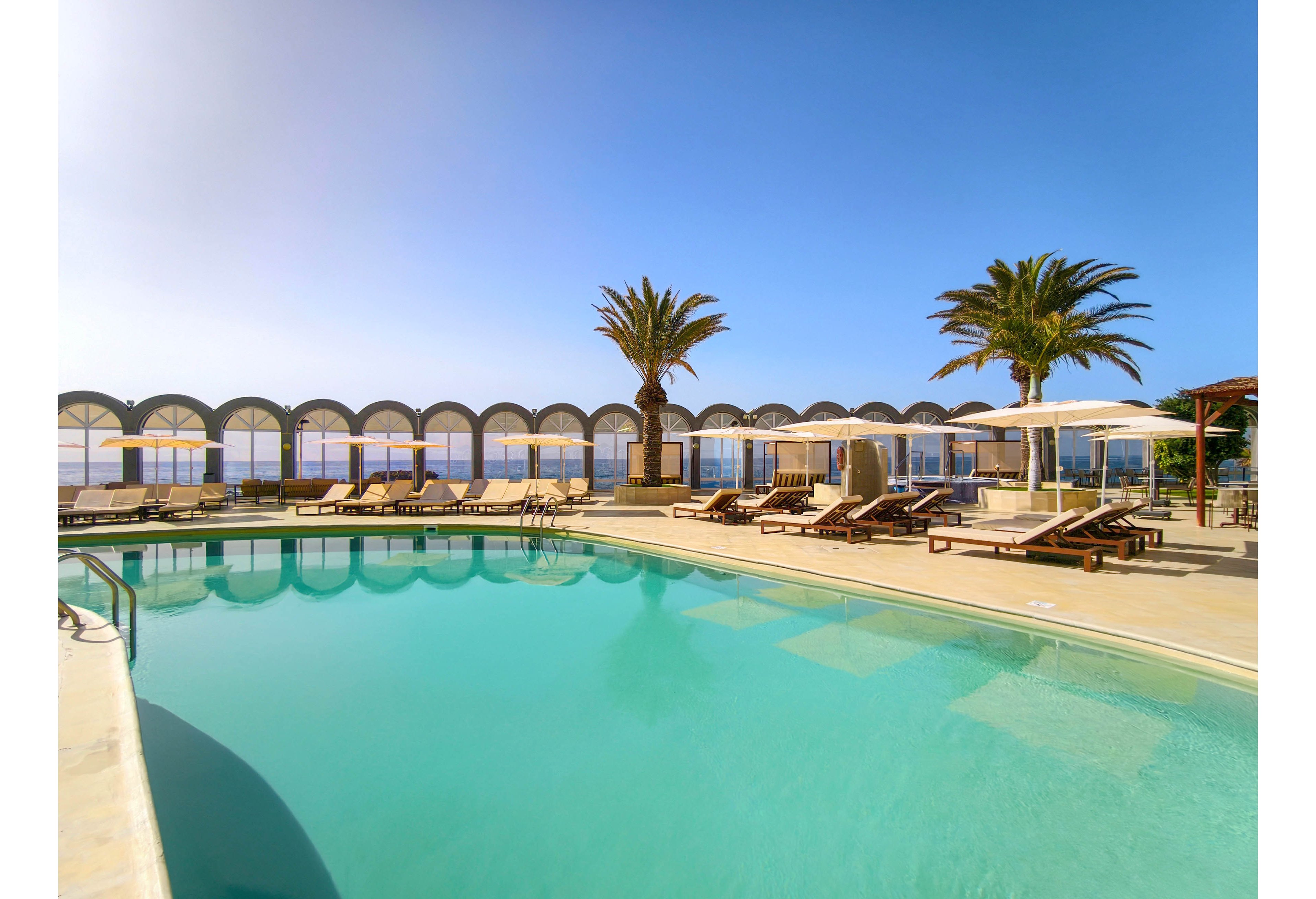 A tranquil outdoor swimming pool sits at a seaside resort under a clear blue sky. Numerous sun loungers and umbrellas line the pool deck, facing the ocean beyond an arched barrier. Palm trees add a tropical touch to the scene, enhancing the vacation atmosphere. The calm water and empty chairs suggest a peaceful, relaxing setting.